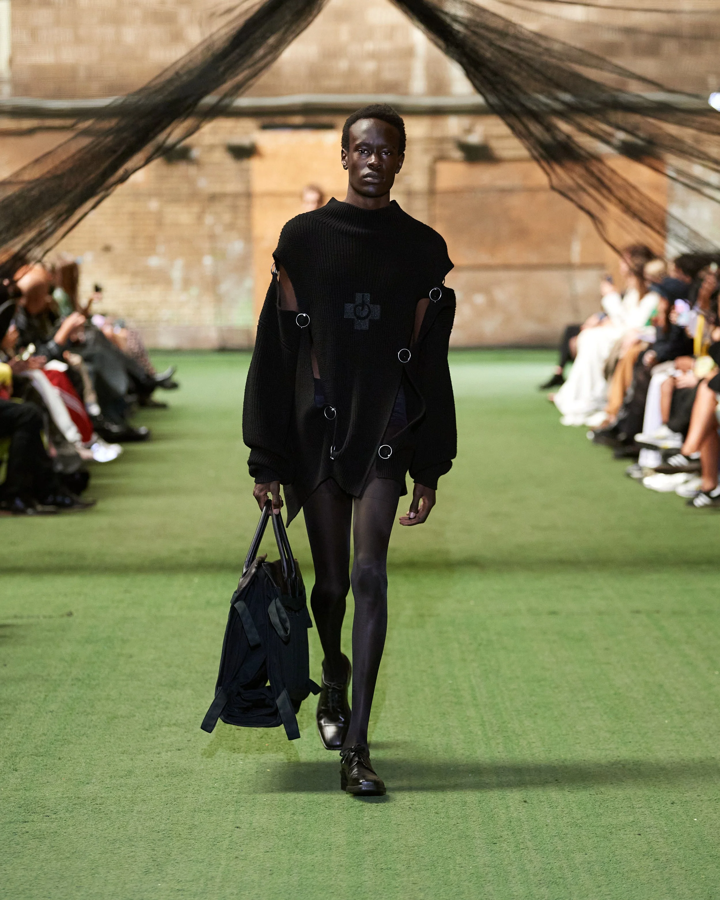 A model walking down a runway at a fashion show, dressed in all black outfit with sheer details, carrying a black bag, with an audience seated on either side of the runway.