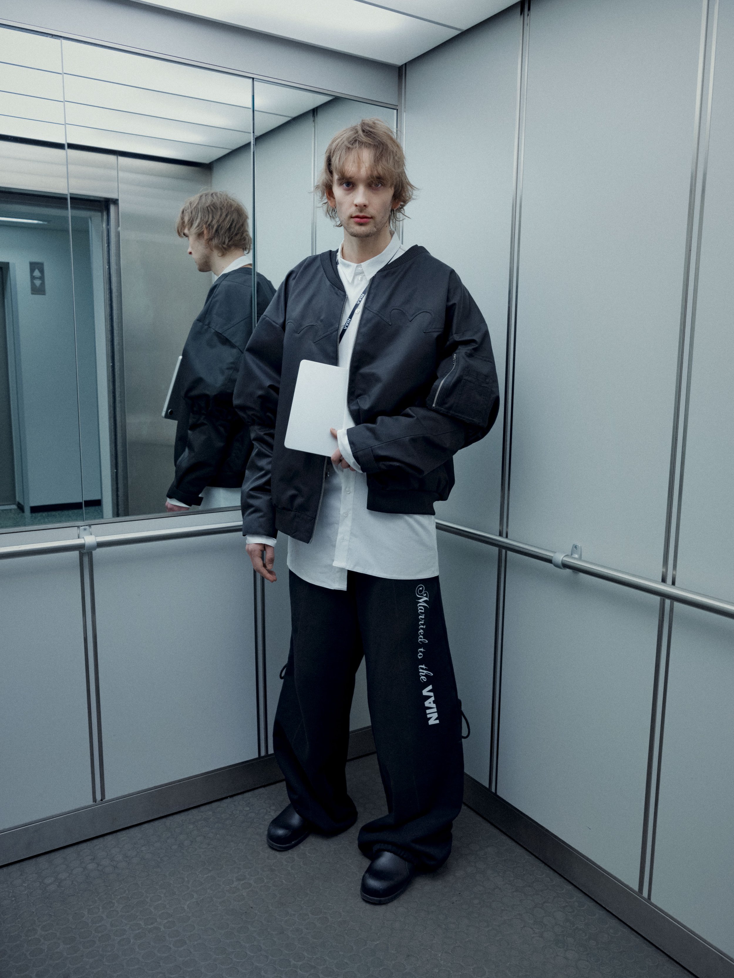 A young man with blonde hair and wearing a black jacket, white shirt, and black sweatpants with writing on the leg, standing in an elevator with a mirror behind him. He is holding a white folder or book.