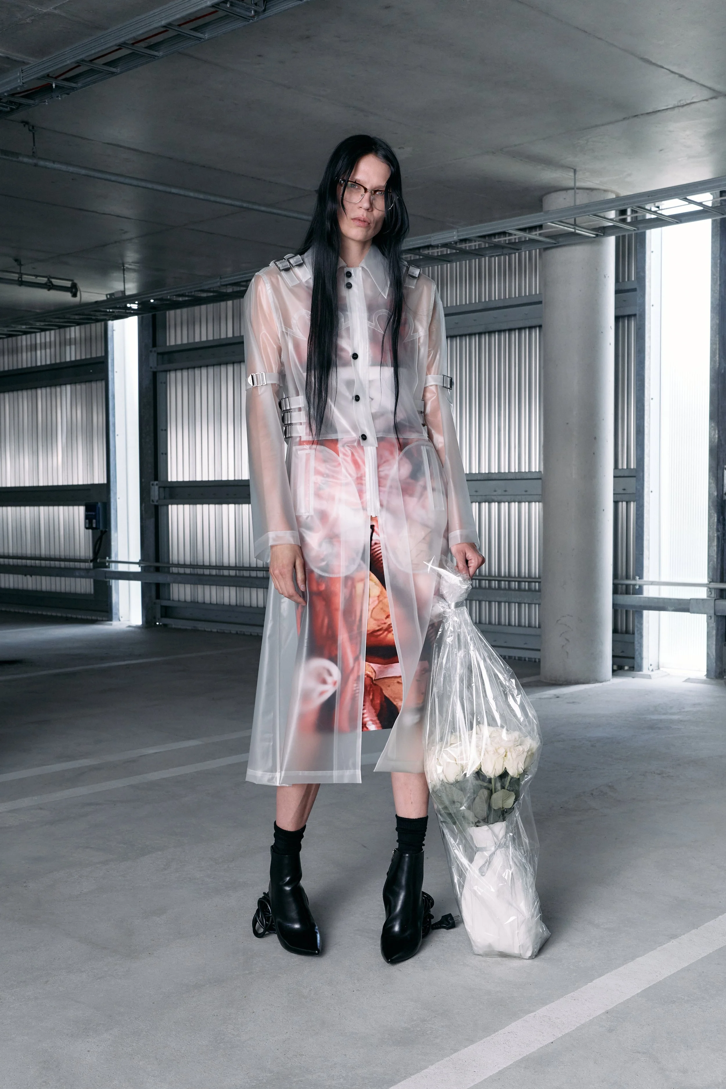 A woman with long black hair and glasses standing in a parking garage, holding a bouquet of white roses in a plastic bag, wearing a transparent coat with fashion prints. VAIN