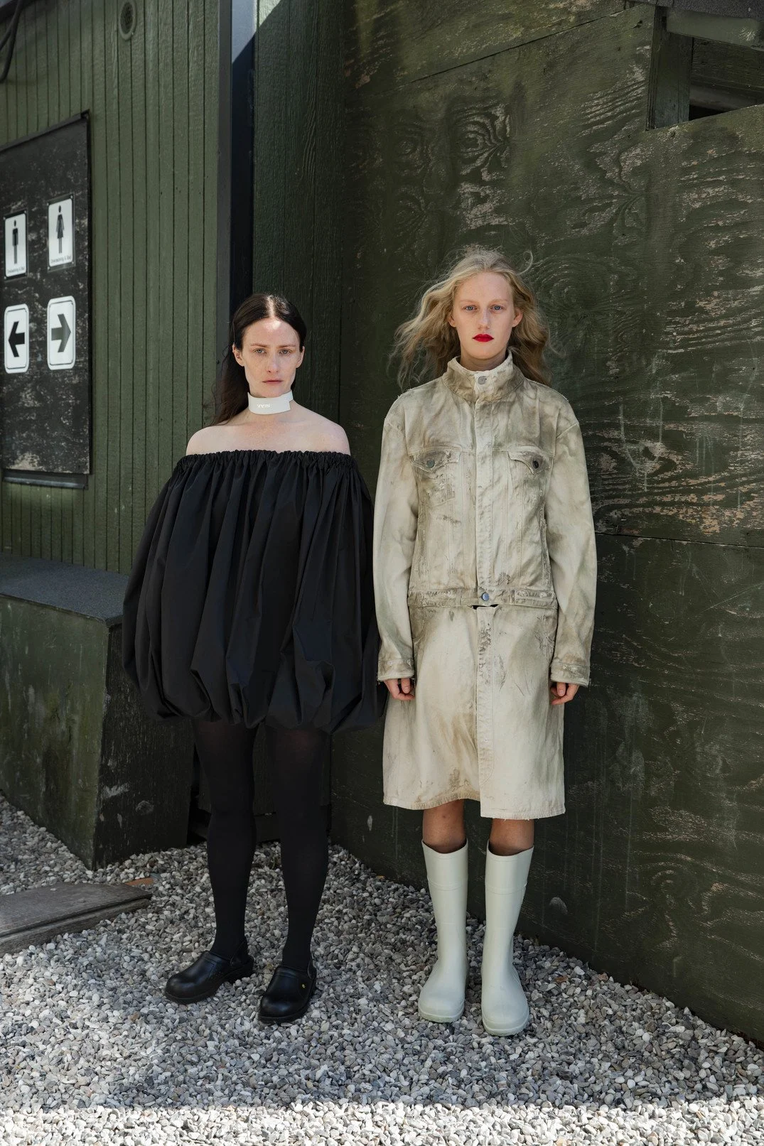 Two women standing outdoors in front of a dark green wooden wall, one in a black off-the-shoulder dress and the other in a worn, beige trench coat with white rain boots.