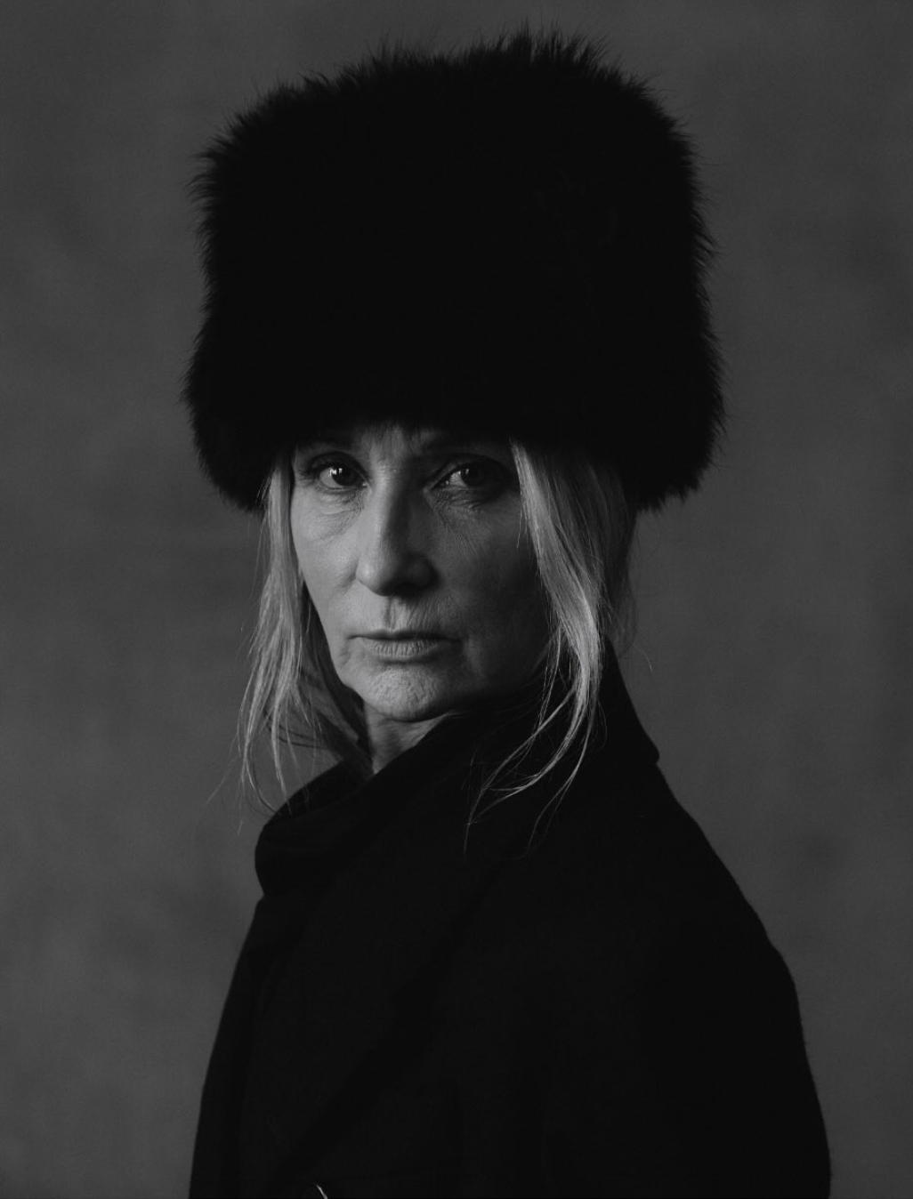 A black-and-white portrait of a woman wearing a large, furry hat and a dark coat, looking directly at the camera with a serious expression.