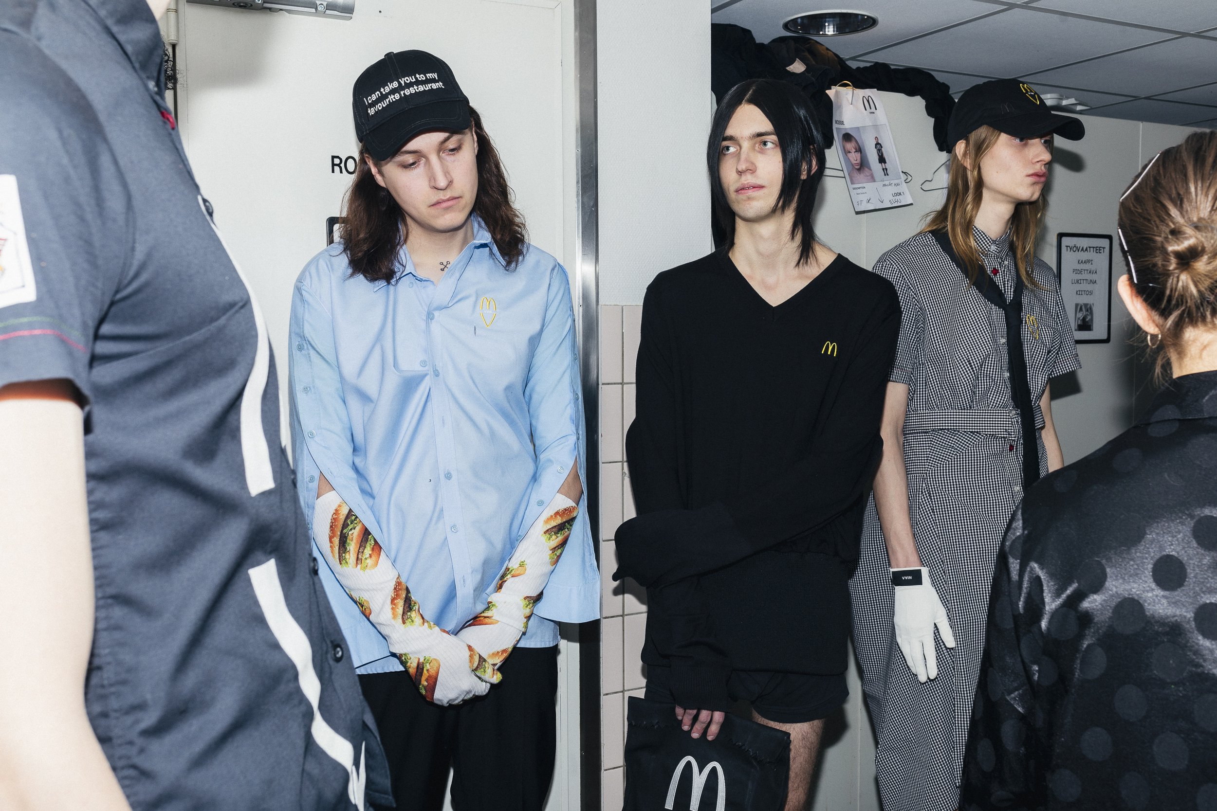 Four young people standing in a line at a fast-food restaurant, wearing McDonald's uniforms and caps, and a woman with her back to the camera talking to them.