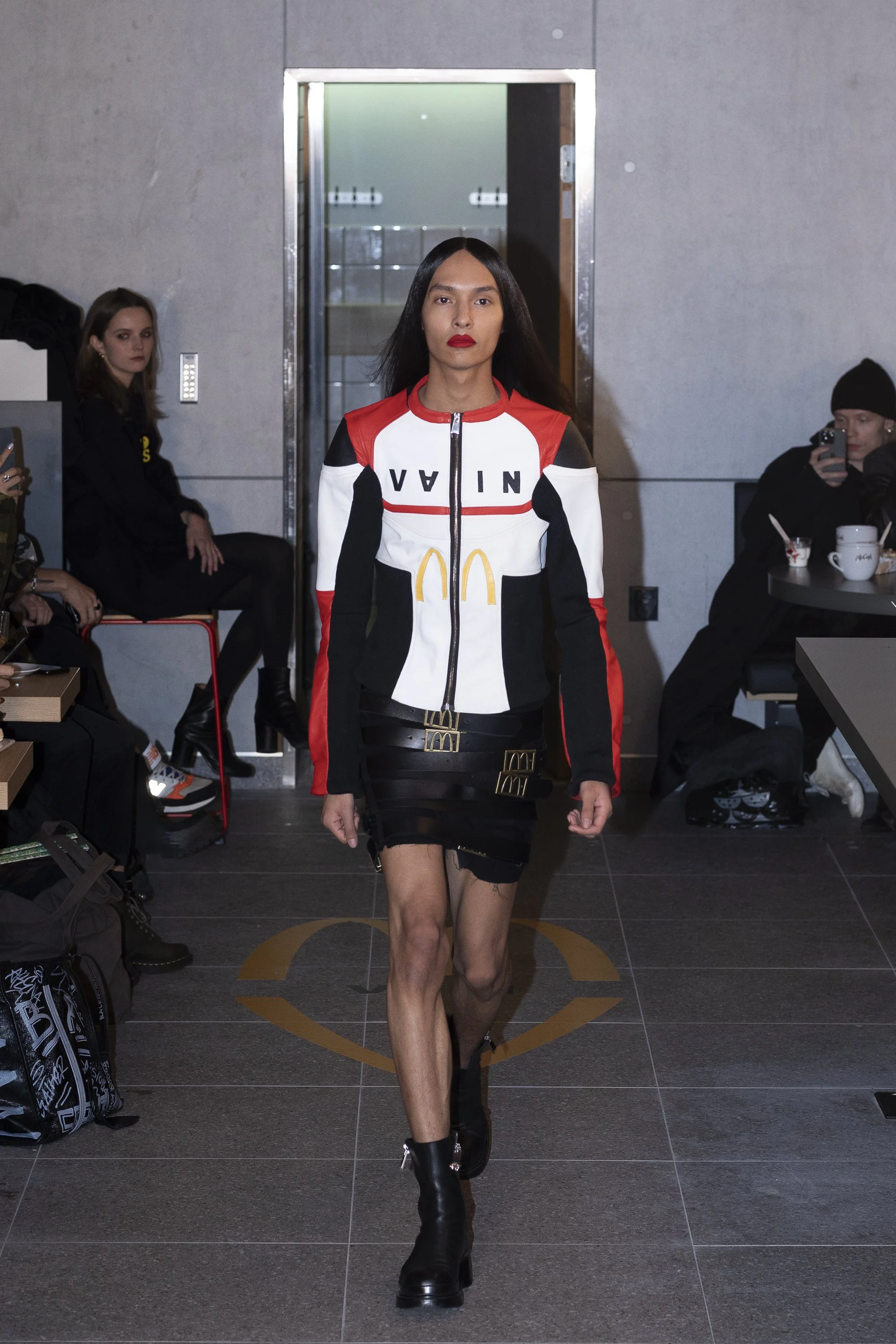 Female model walking on runway in a McDonald's-themed jacket, black skirt, and black boots, with audience seated on either side. VAIN x Mcdonald's