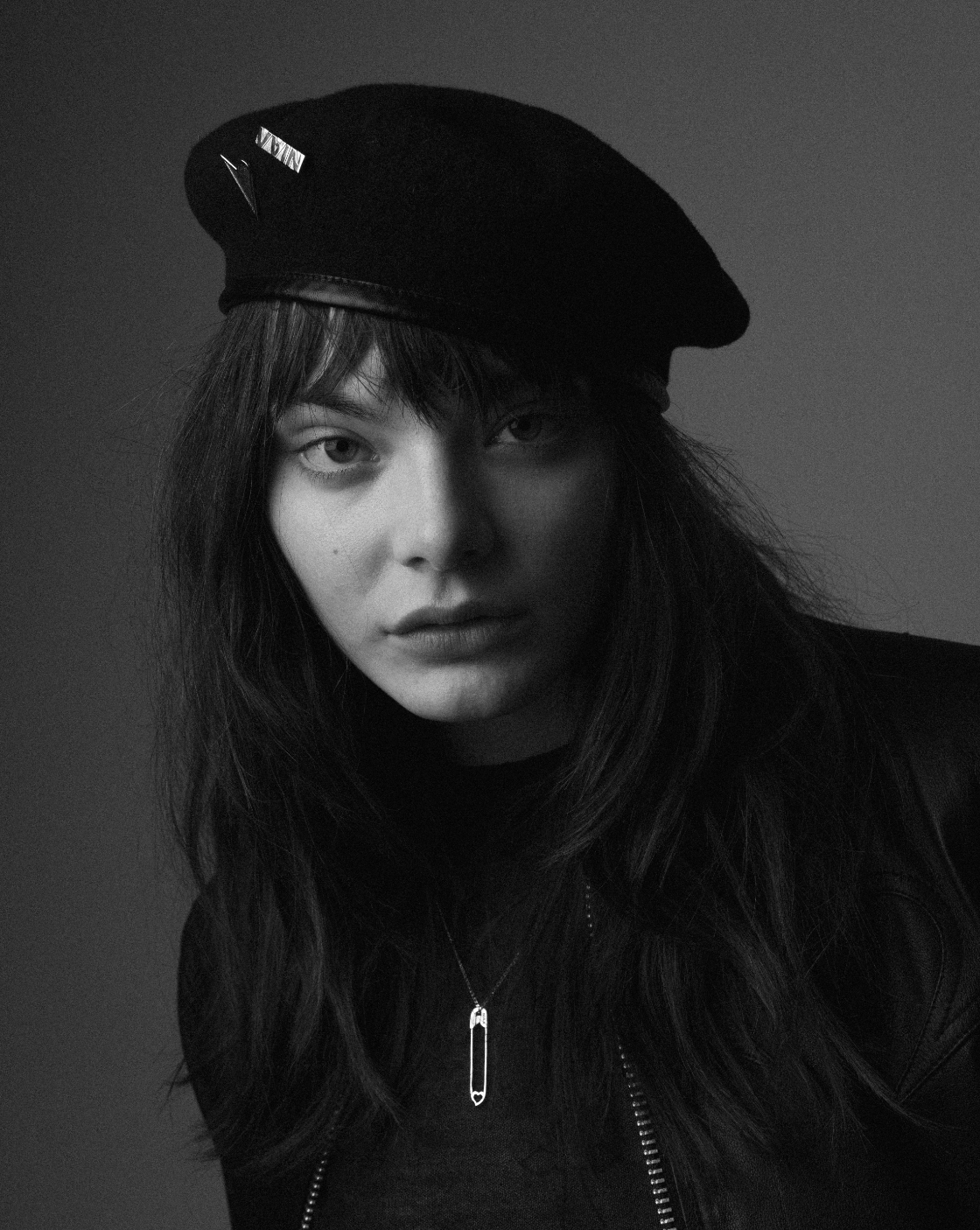Black and white portrait of a woman wearing a beanie with patches, leather jacket, and a necklace with a safety pin pendant.