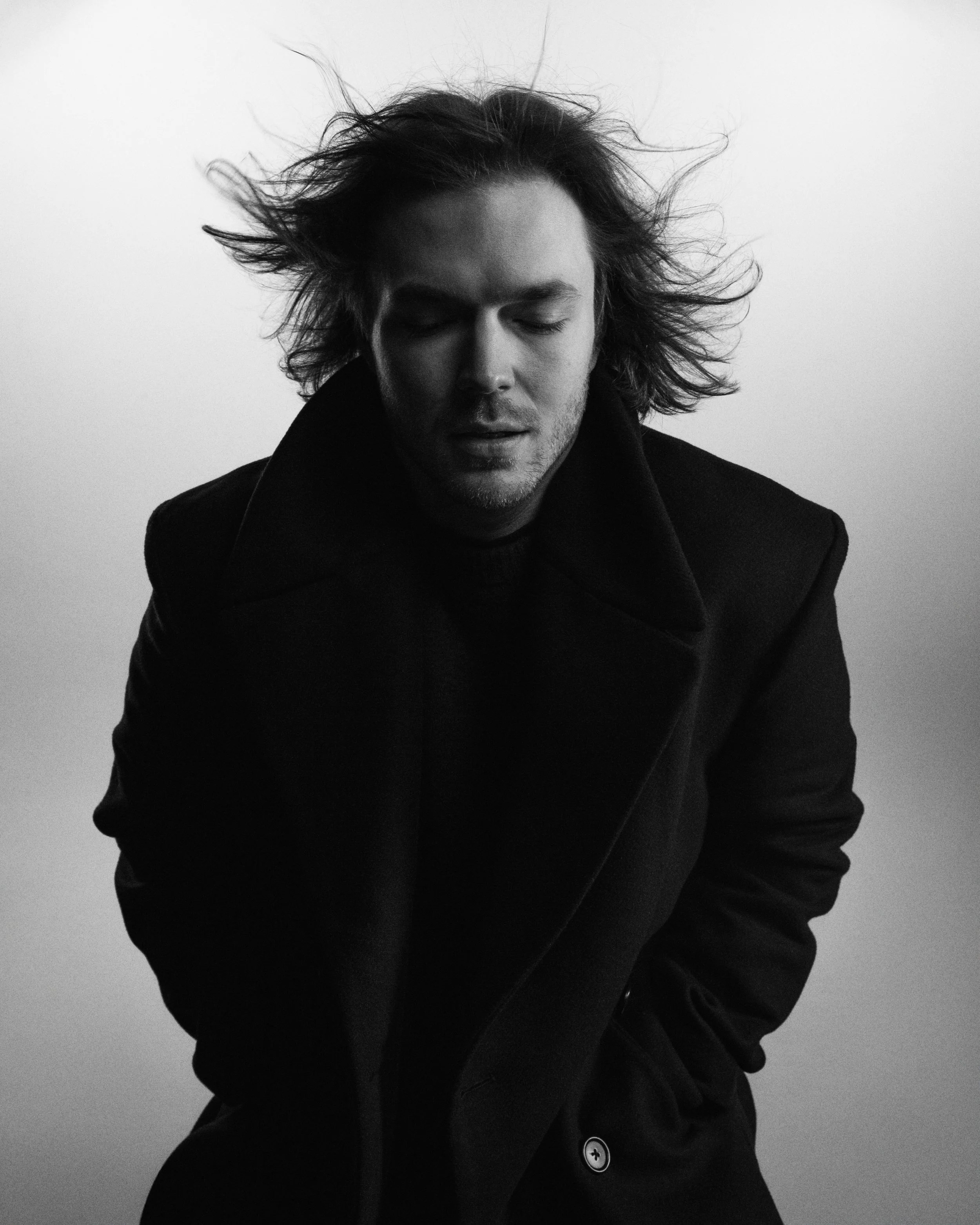 Black and white portrait of a man with long, tousled hair, eyes closed, wearing a dark coat against a plain light background. Jaakko Kulta. tupakkaa ja rakkautta