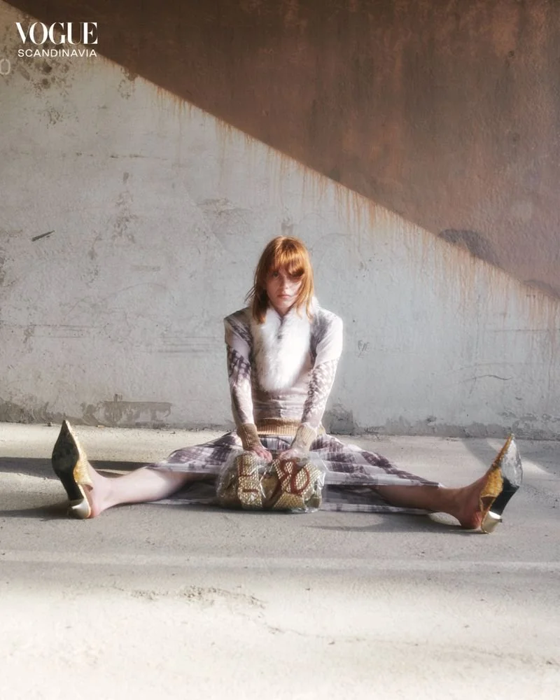 A woman with red hair and a patterned dress sitting on the floor with her legs spread wide apart, holding a bag in front of her. The setting appears to be an industrial or unfinished space with a textured wall and concrete floor. Vogue Scandinavia