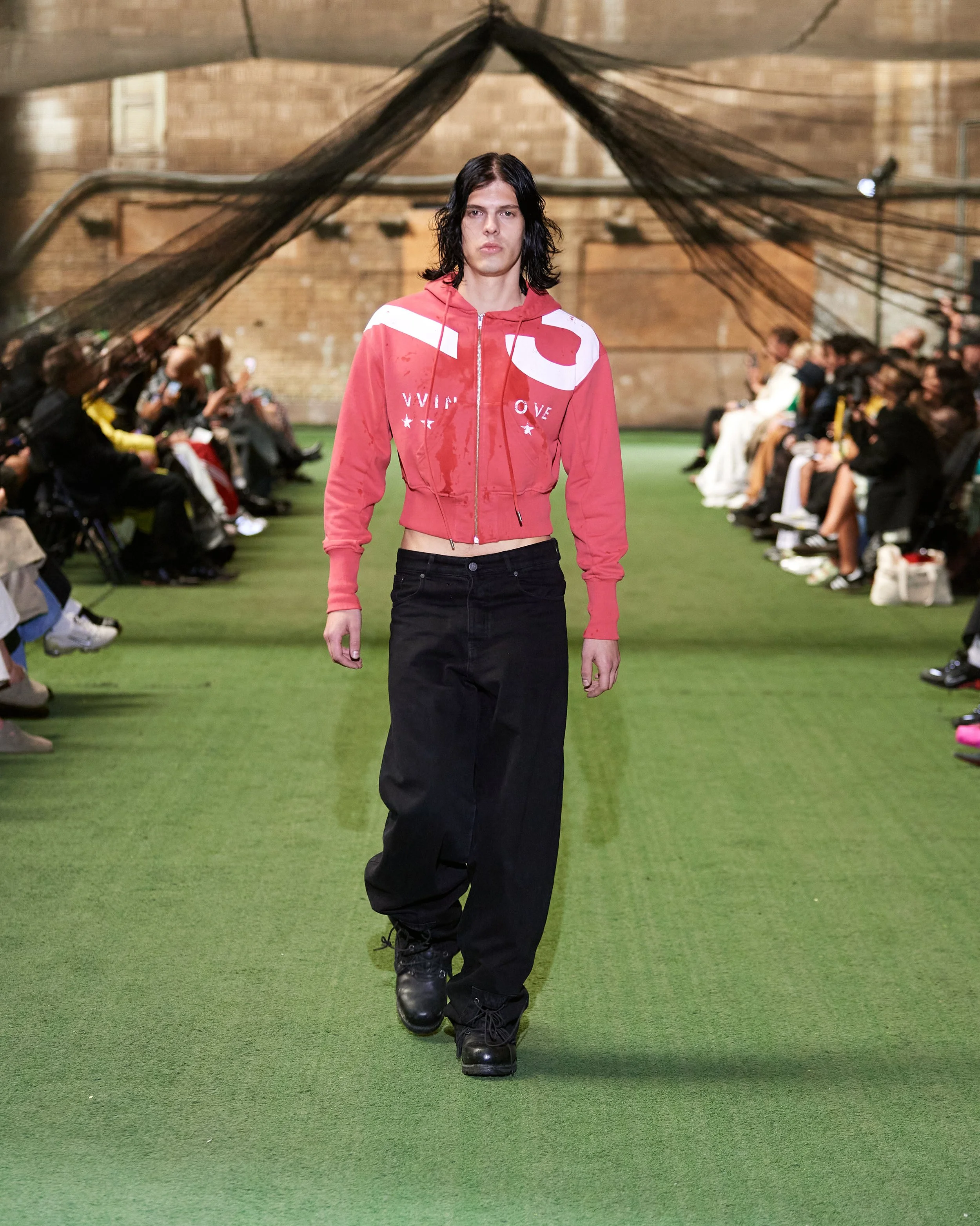 Model walking down a green runway at a fashion show, wearing a red cropped zip-up hoodie with white graphics, black loose-fitting pants, and black boots, audience seated on either side.