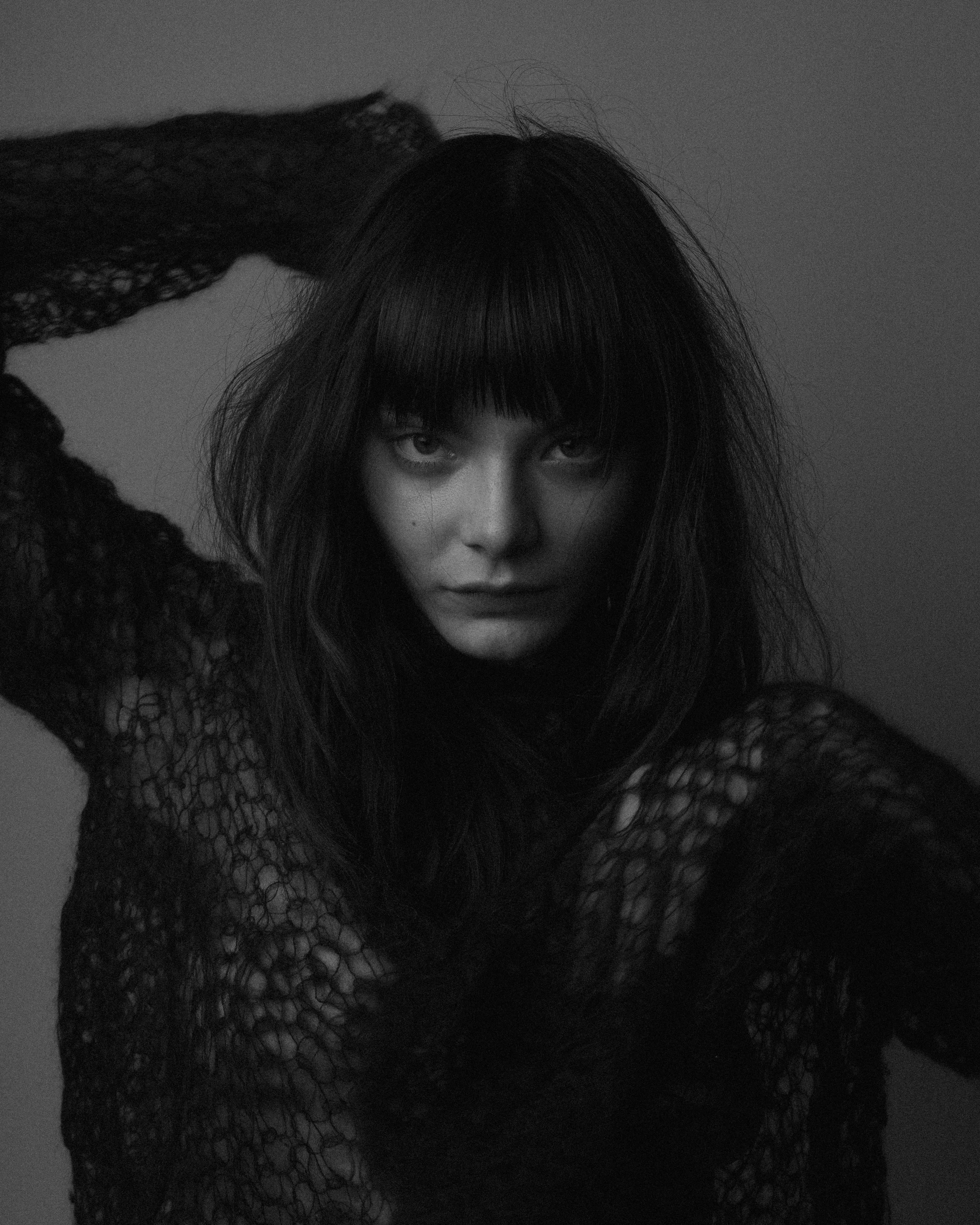 Black and white portrait of a woman with long, messy hair, wearing a lace top, gazing directly at the camera with her hand behind her head.