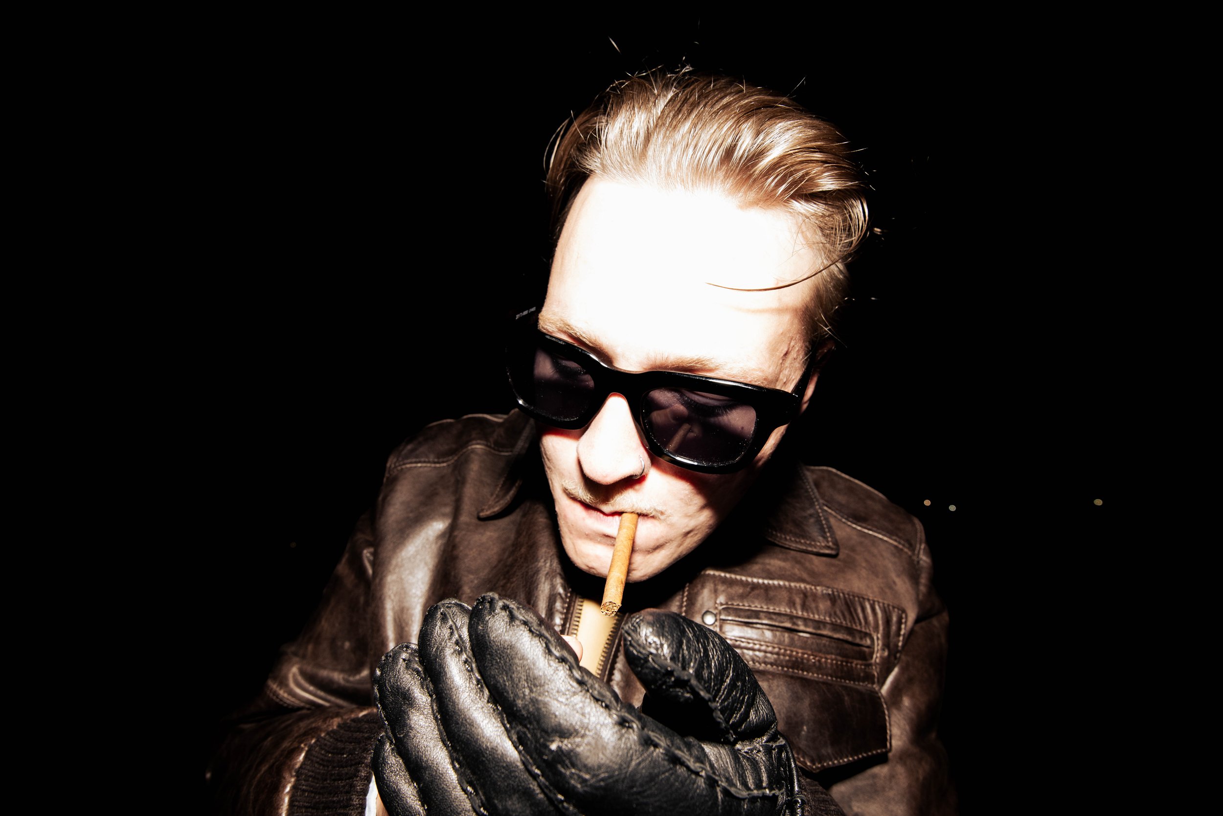 A person with blonde hair wearing black sunglasses and a black leather glove, lighting a cigarette with a lighter at night. 6kulma! kill your idol