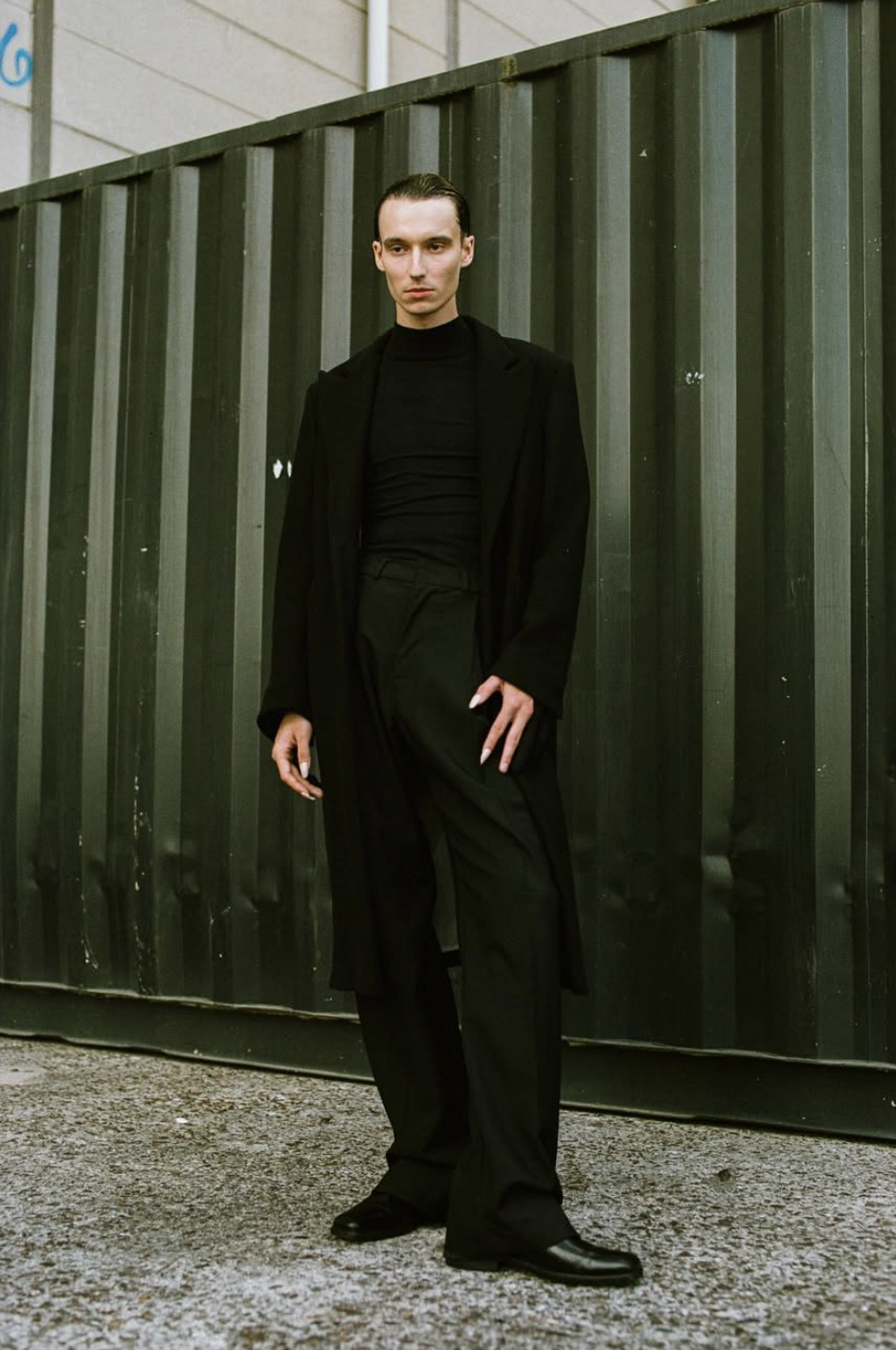 A young man stands against a green metal fence, dressed in all black, wearing a long blazer, turtleneck shirt, dress pants, and shoes, with slicked-back hair. verner vuorela. VAIN. cphfw