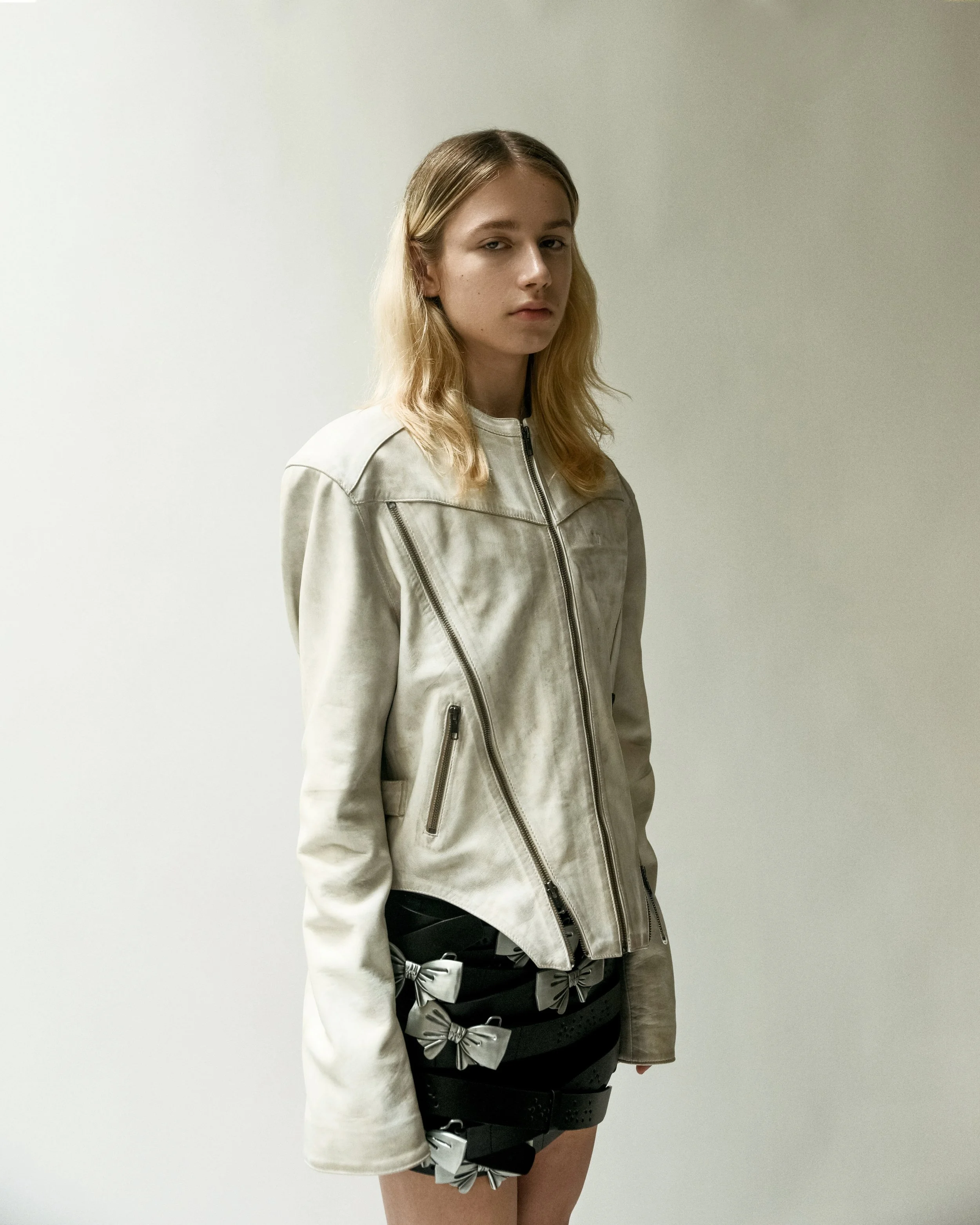 Young woman with blonde hair wearing a light-colored leather jacket and a black skirt with large gray bows, standing against a plain white background.