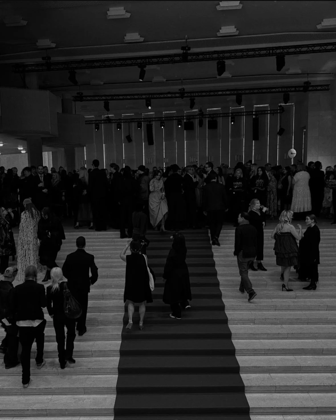 People attending a formal event or celebration indoors. Some are on a staircase leading to a stage or higher area, others are mingling in groups across the room. The scene is in black and white. Finnish fashion awards