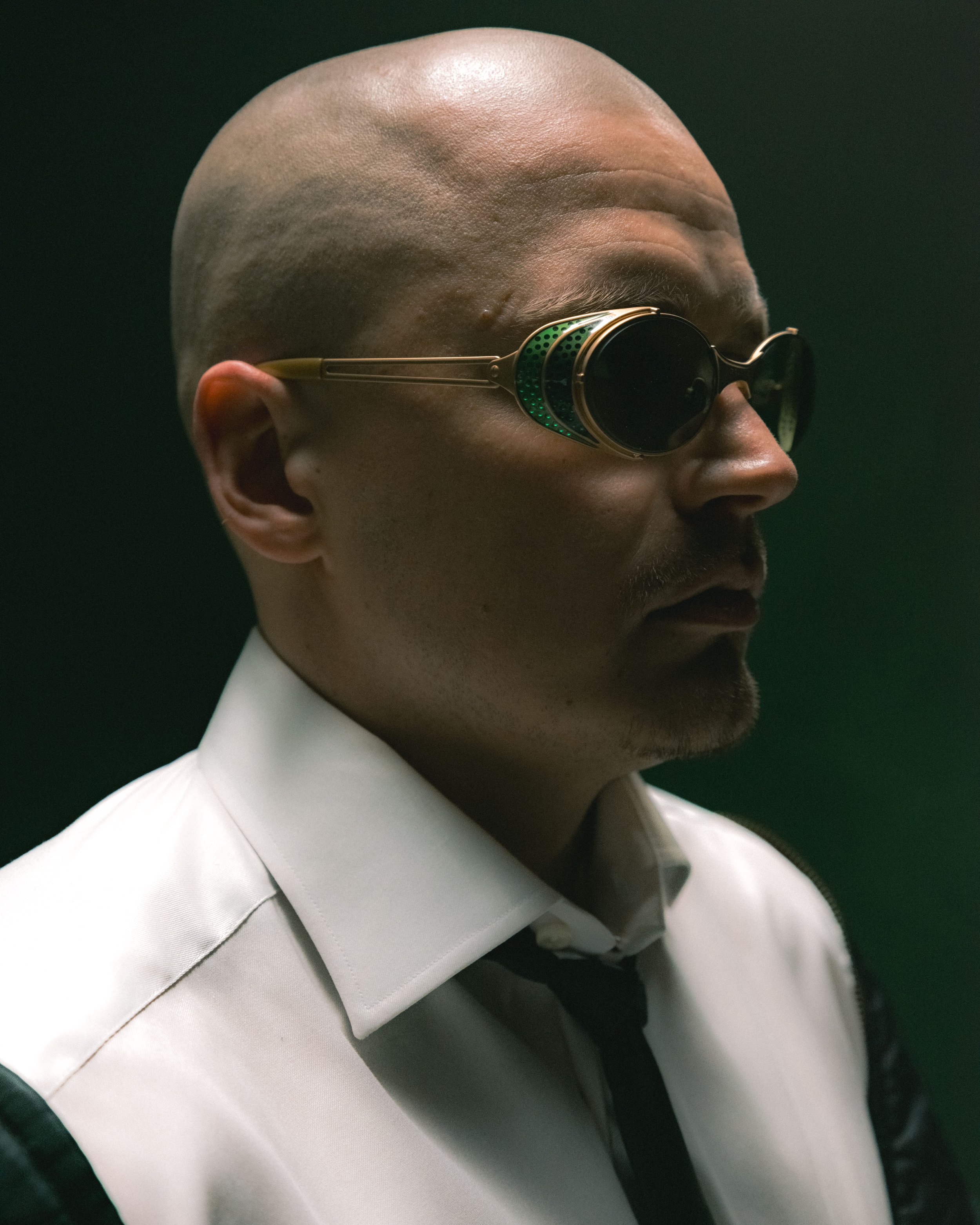 A man with a shaved head wearing futuristic sunglasses, a white dress shirt, and a black tie, against a dark background. gettomasa sexmane