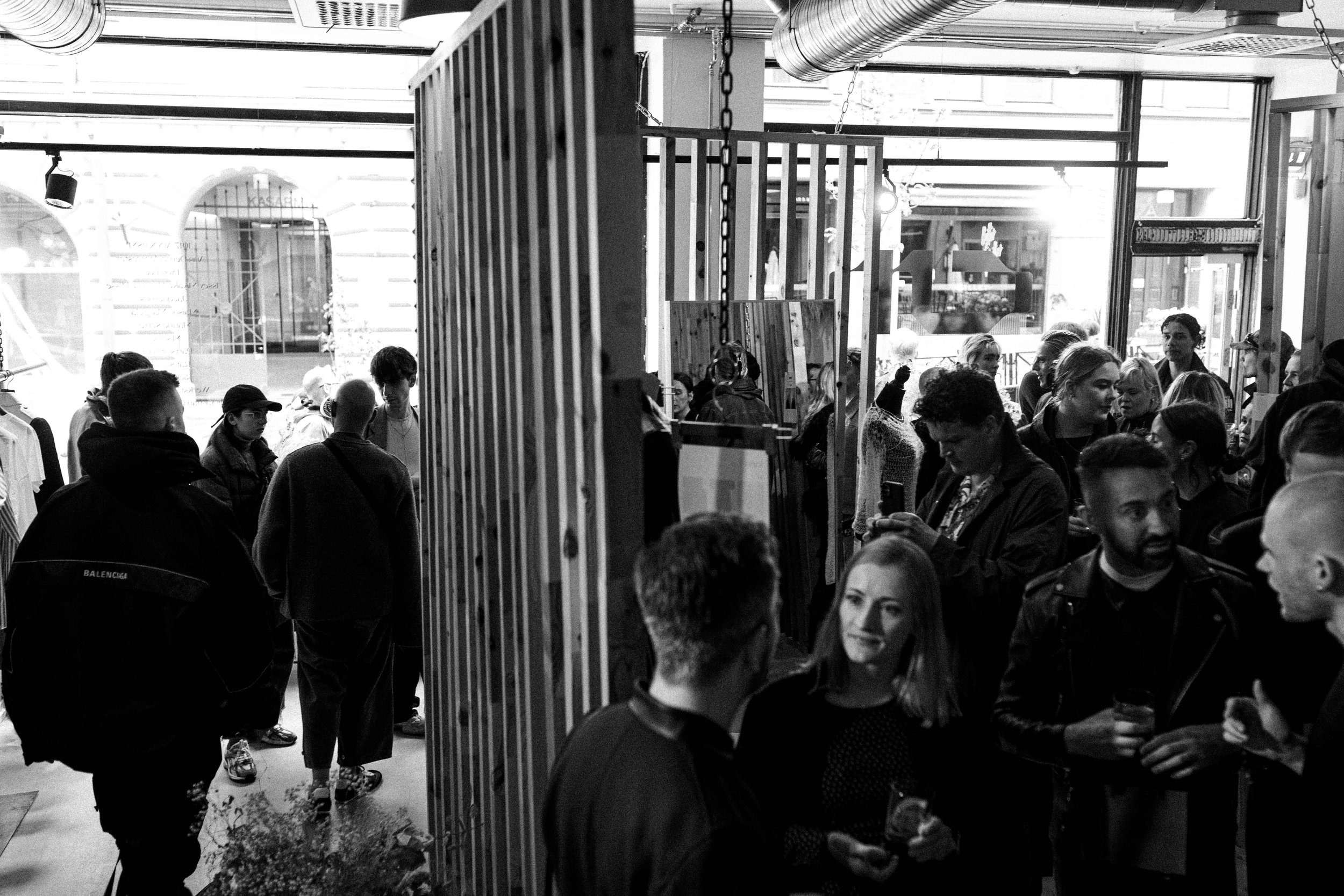 People gathered inside a retail store, some engaging in conversation while others browse or use their phones, with clothing displays and large windows in the background.