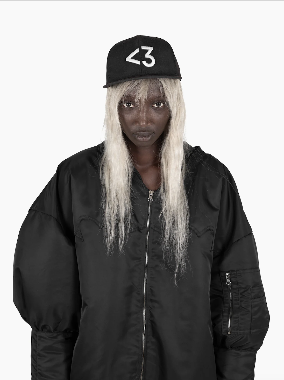 A person with long platinum blonde hair wearing a black baseball cap with white text, a black bomber jacket, and posing against a plain white background. VAIN, jimivain