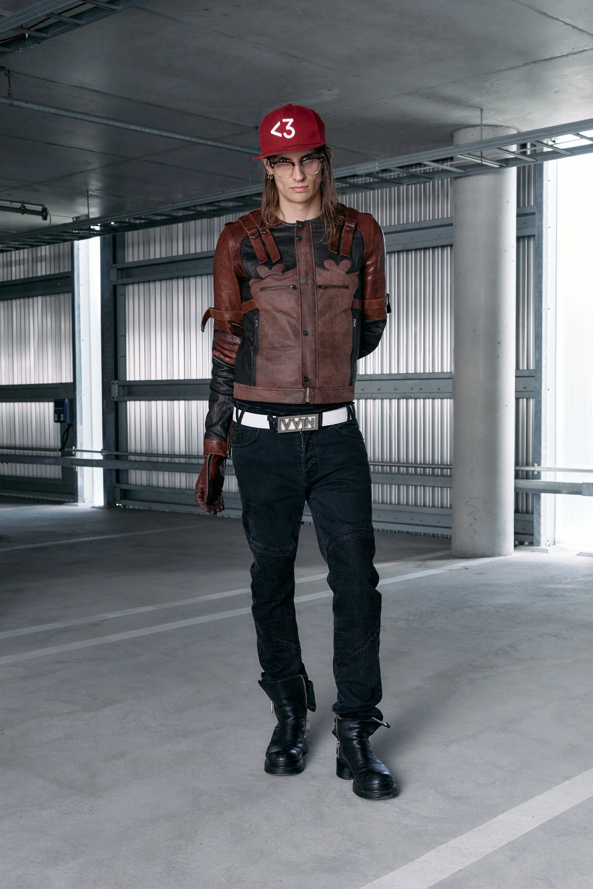 A young woman stands in a parking garage wearing a brown and black leather jacket, black pants, combat boots, a red cap with a white symbol, and glasses. vain
