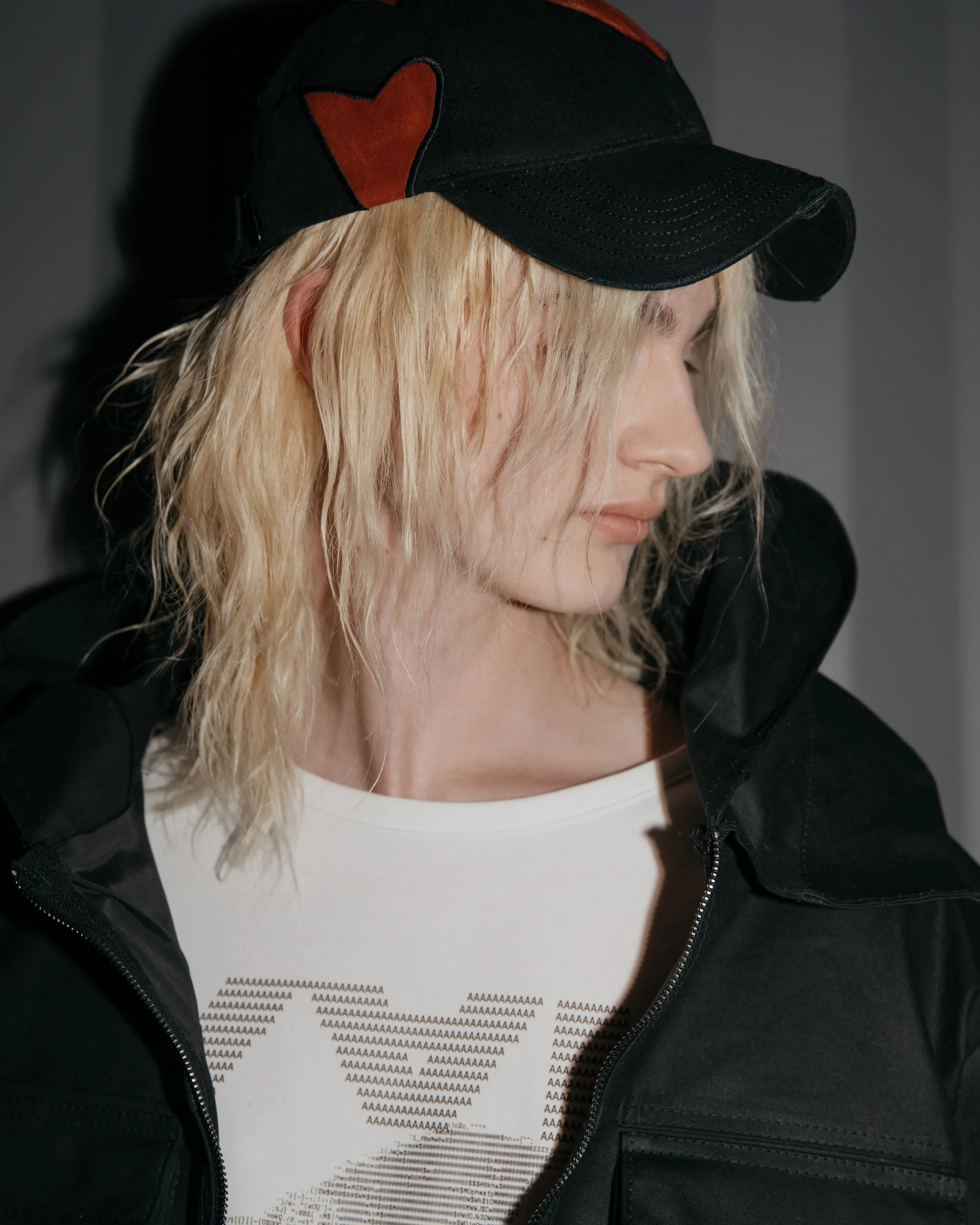 A woman with blond hair wearing a black cap with a red heart, a white t-shirt with a black graphic, and a black jacket, looking downward with her profile visible. vain cphfw