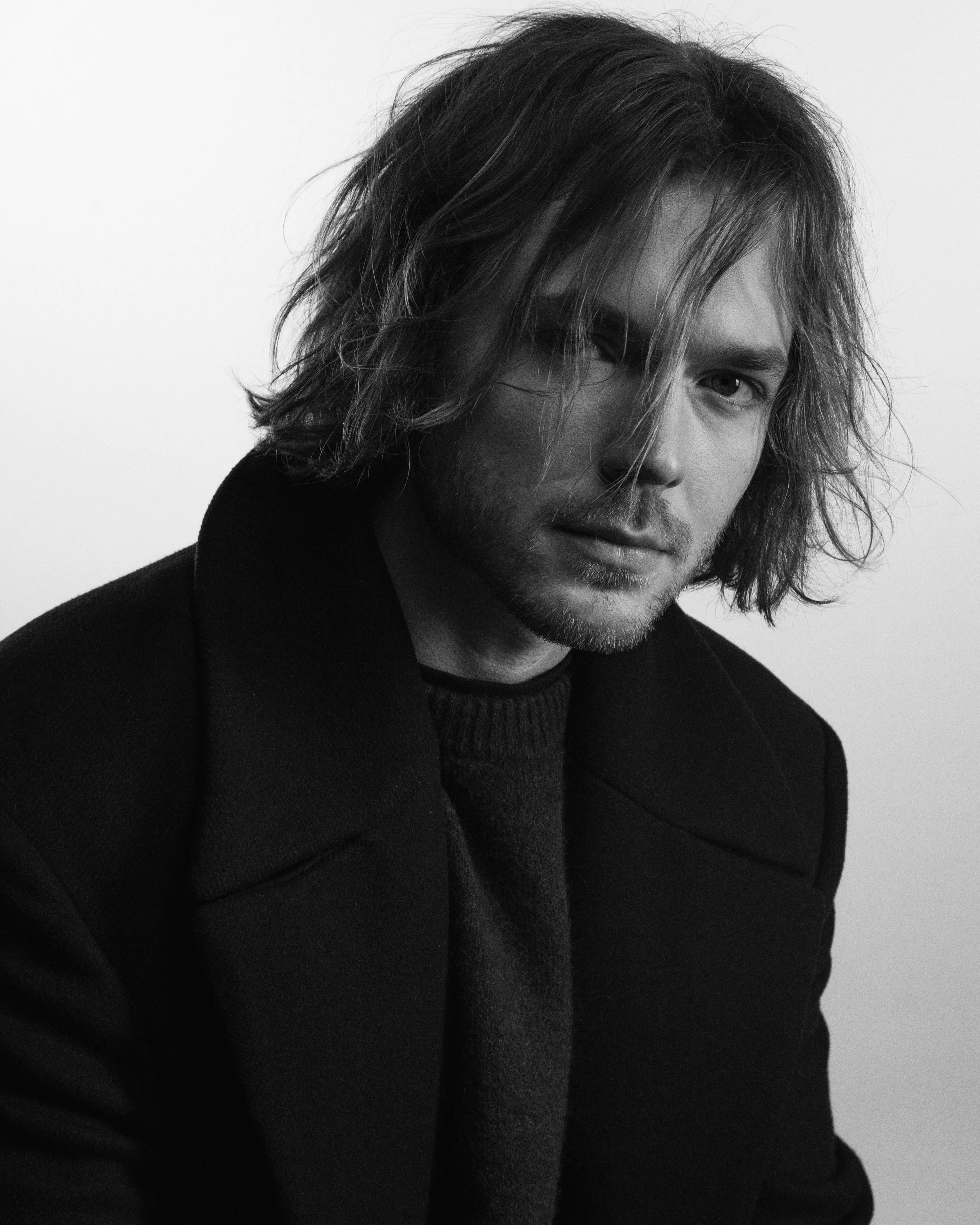 A black and white portrait of a man with shoulder-length wavy hair, wearing a dark coat and a sweater, looking at the camera with a slight serious expression. jaakko kulta 