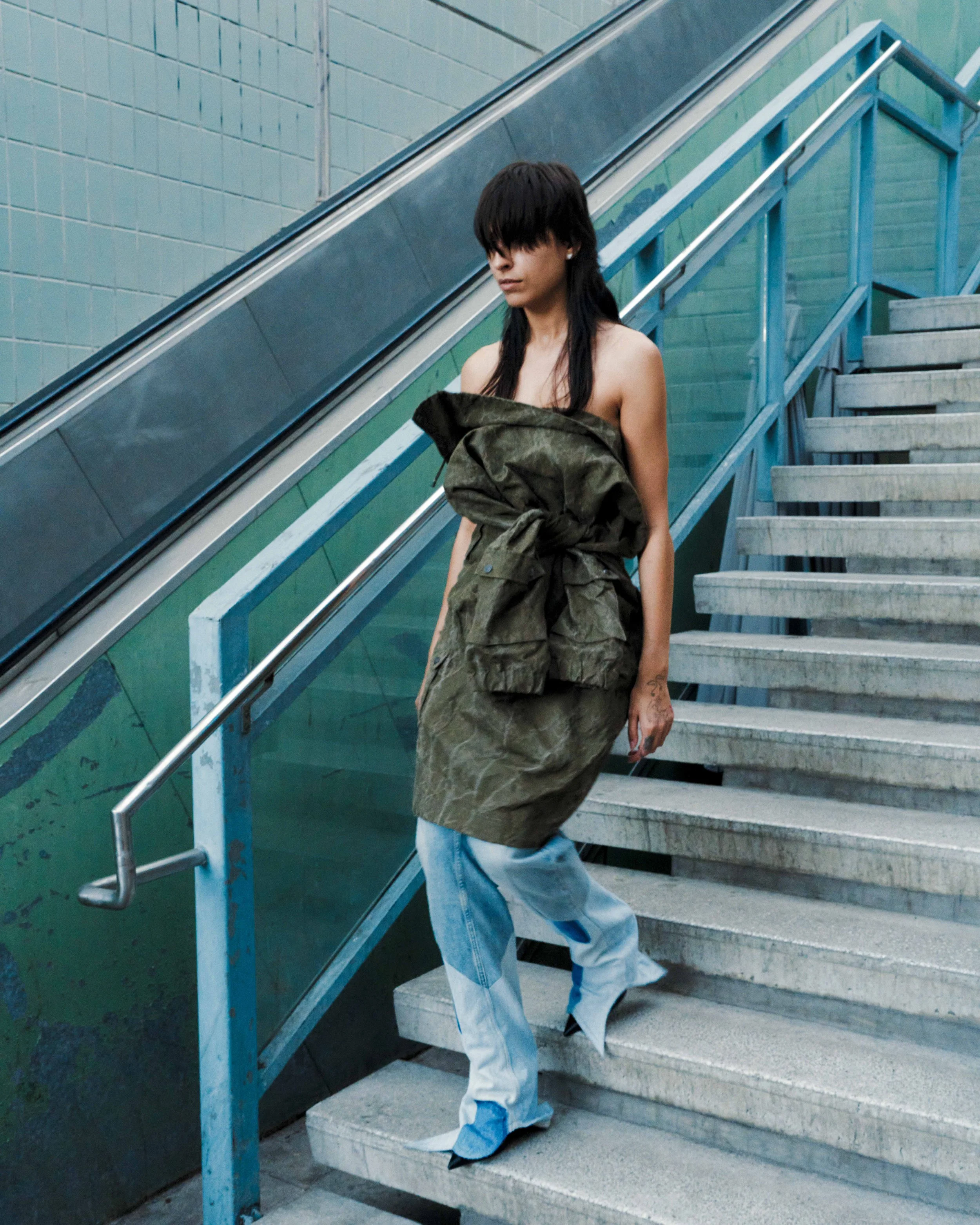 A person with long dark hair descending outdoor concrete stairs, wearing baggy blue pants and an oversized green jacket draped over their shoulders, against a background of turquoise tiled wall and metal railing. VAIN fashion