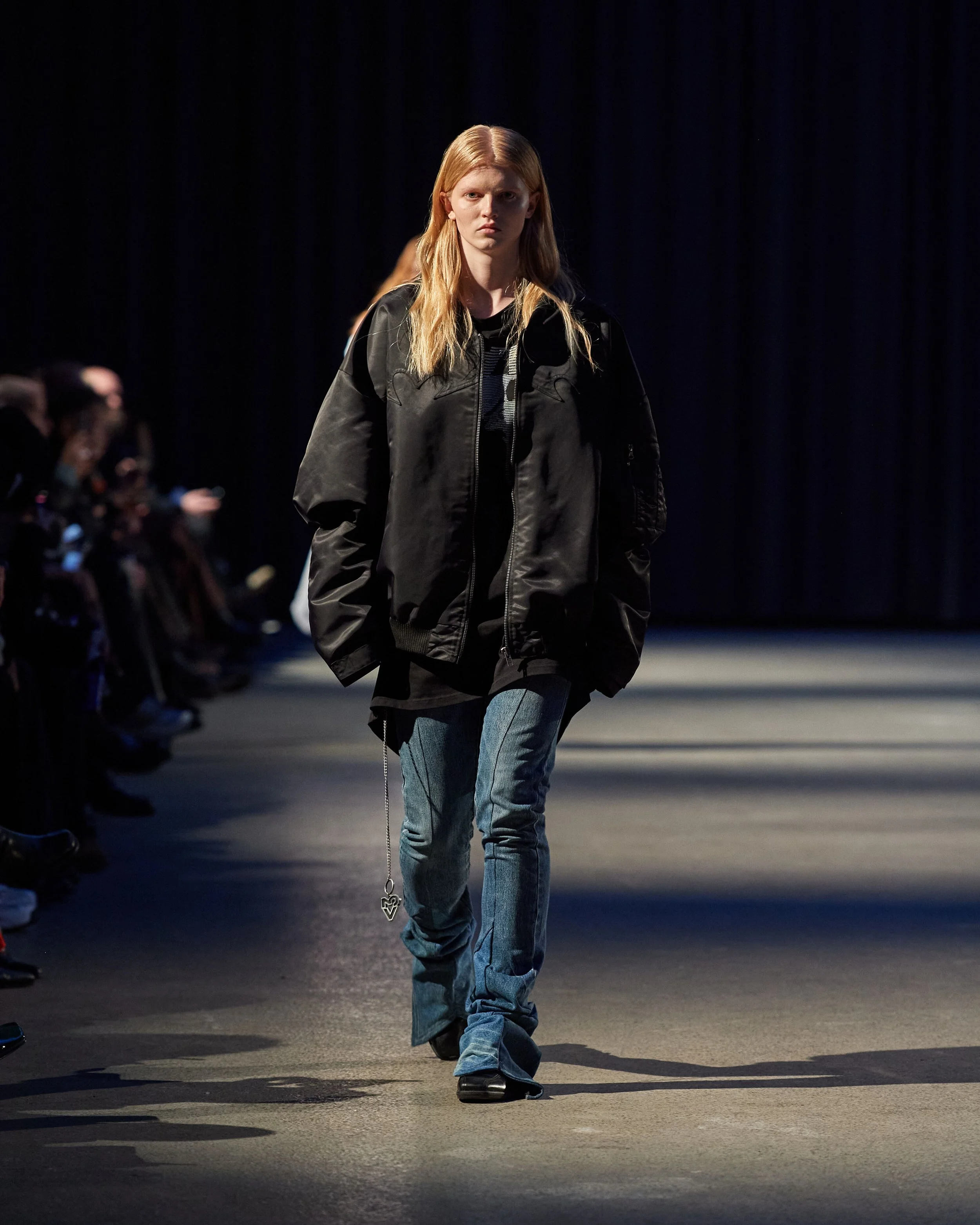 A female model walking down a runway during a fashion show, wearing a black oversized jacket, black shirt, and blue jeans, with audience members seated on either side. VAIN cphfw