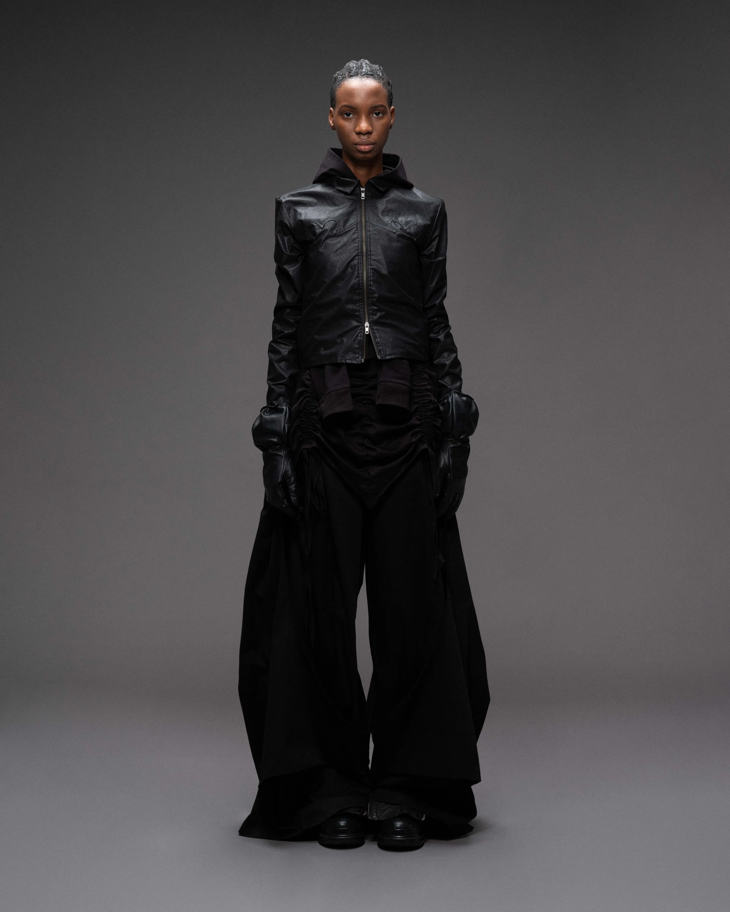 A woman standing in a studio with a gray background, wearing an all-black outfit including a leather jacket, wide-leg pants, gloves, and black shoes. VAIN