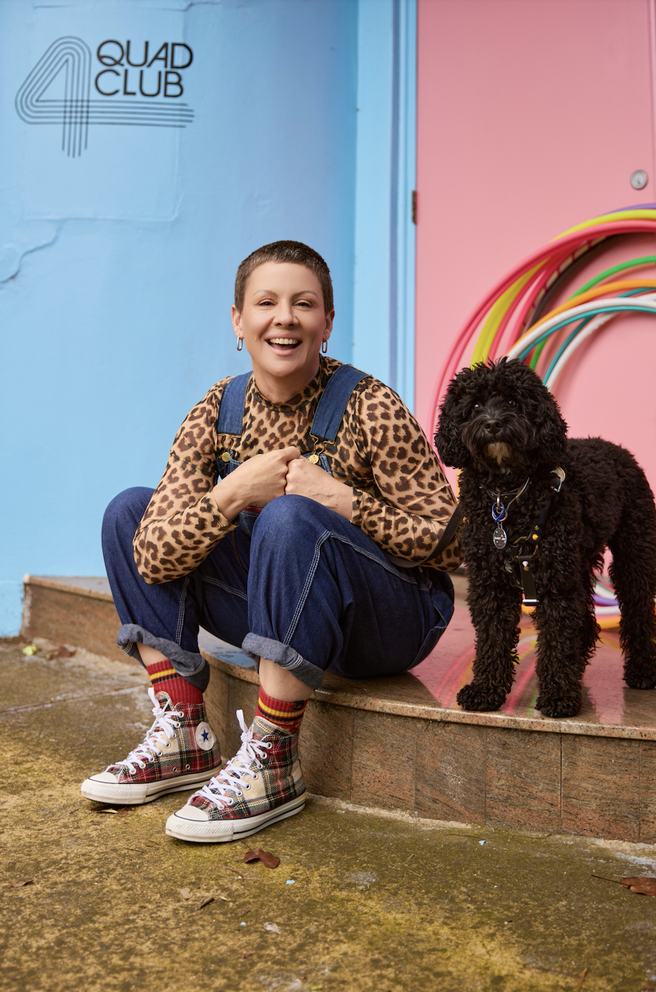 Queer woman with short hair and leopard top, blue overalls, plaid high-top sneakers, smiling and sitting on a step with a black Cavoodle beside her, outdoors in front of a wall with the words 'QUAD CLUB' and rainbow hula hoops in Melbourne.