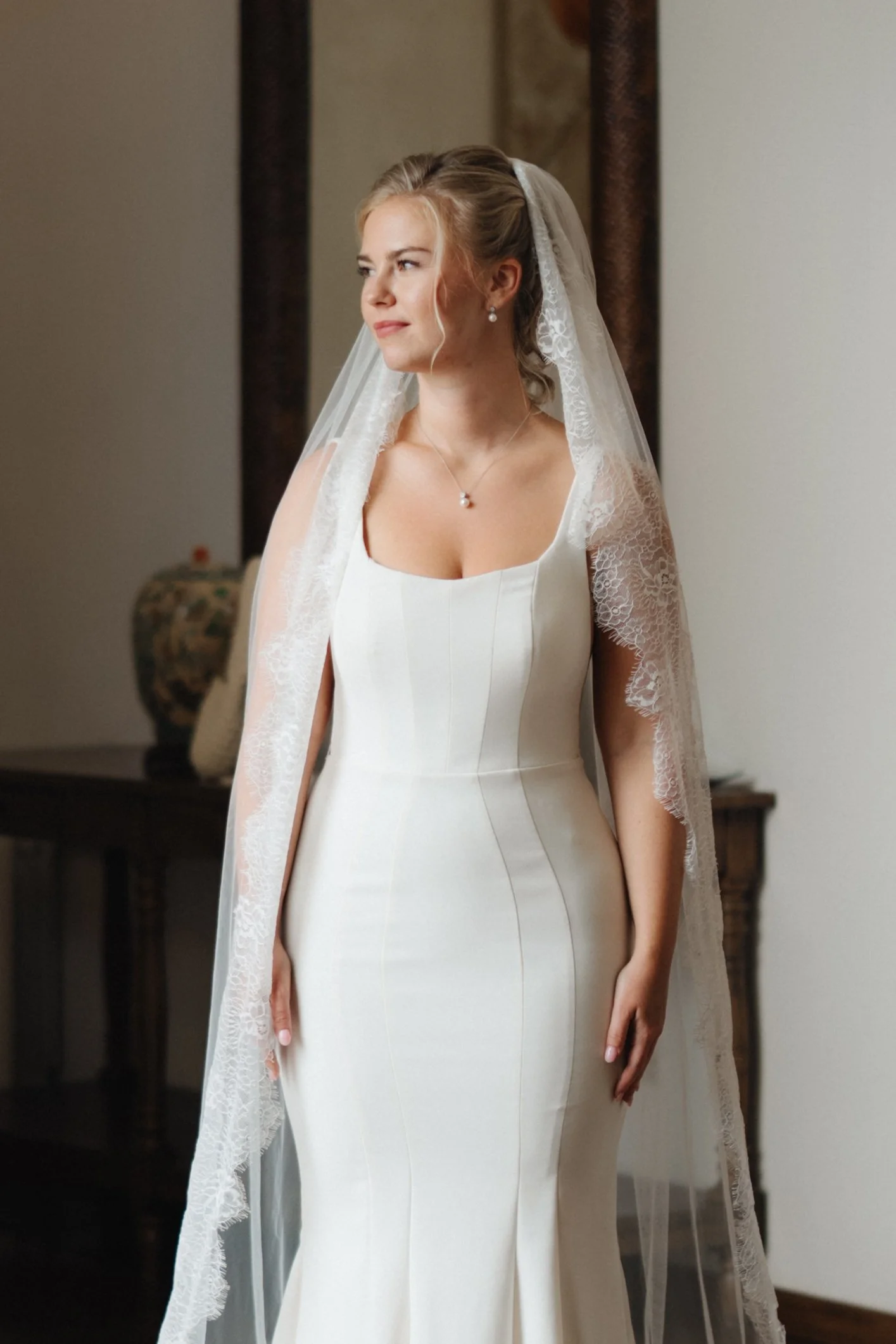 Mujer vistiendo vestido de novia con rostro de perfil, fondo de interior con jarrón decorativo.