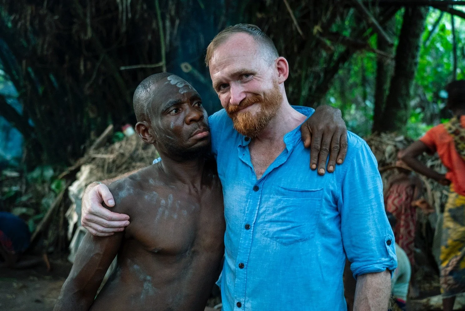 Two men, one shirtless with dark skin and traditional markings, and another with red hair and beard wearing a blue shirt, embrace each other in a lush, green jungle setting.
