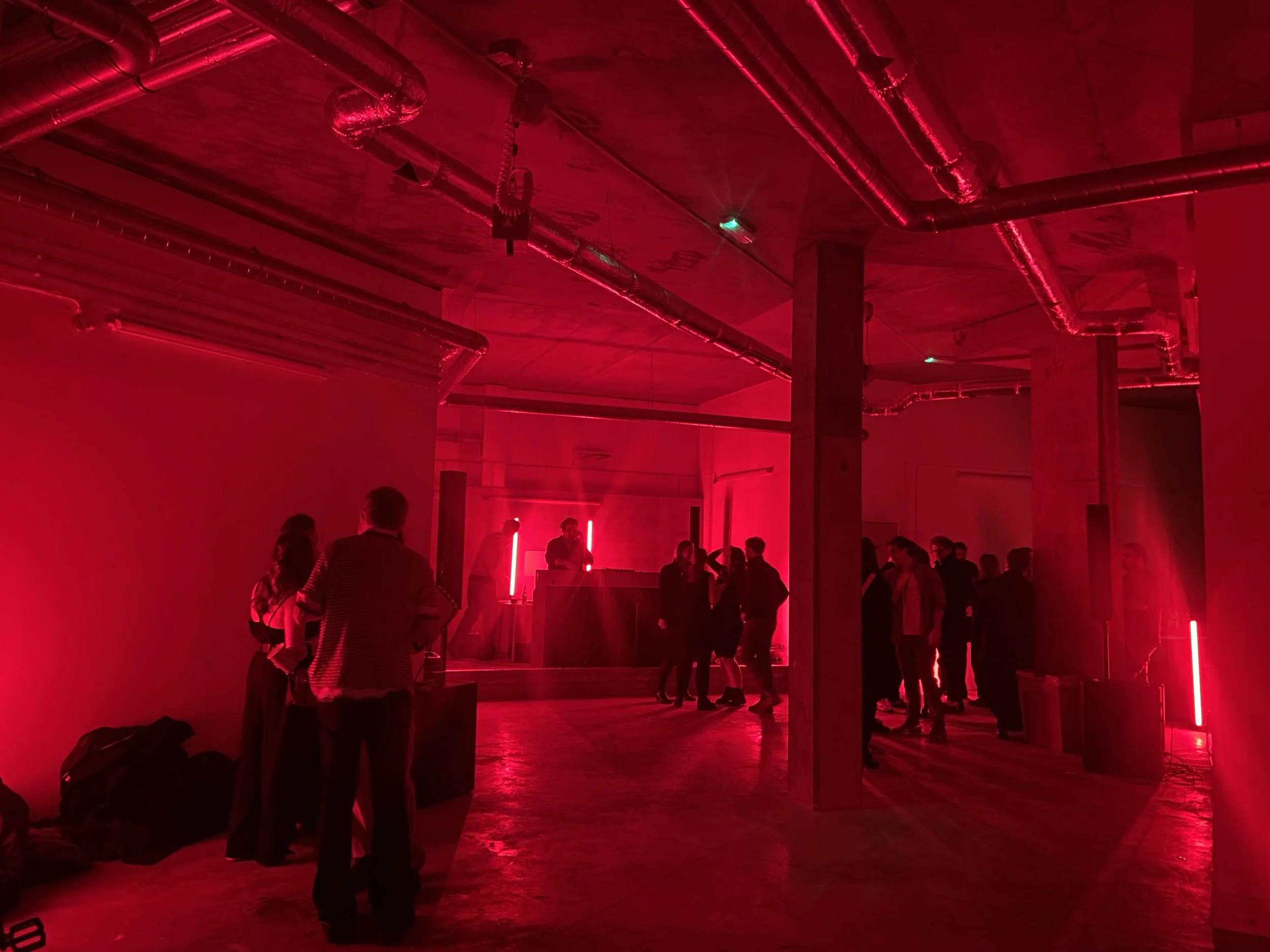 People gathering in a room with red lighting, industrial-style ceiling with visible pipes, and some standing near a DJ booth.