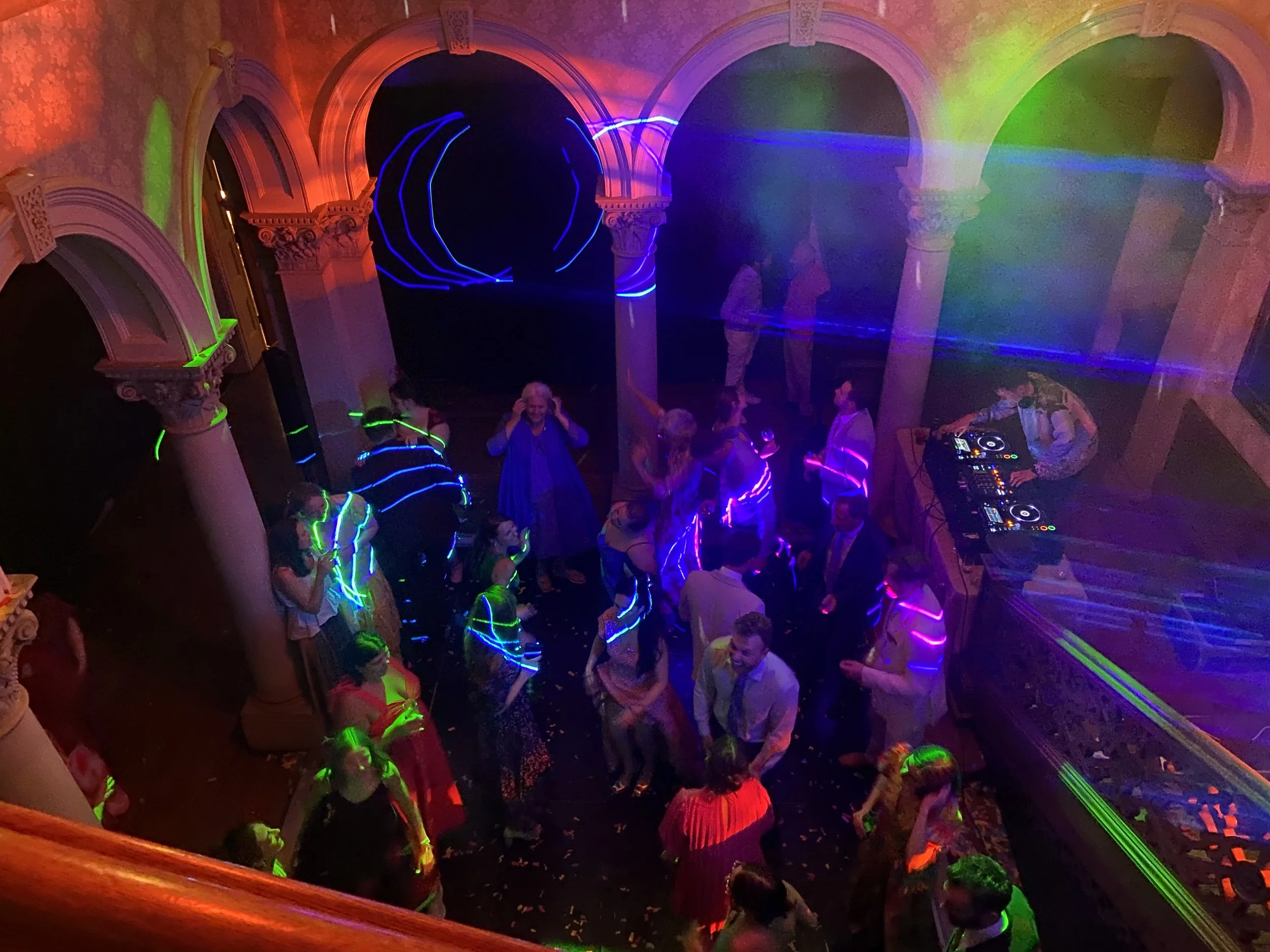 People dancing and enjoying music in a dimly lit club with colorful laser lights and an ornate balcony.