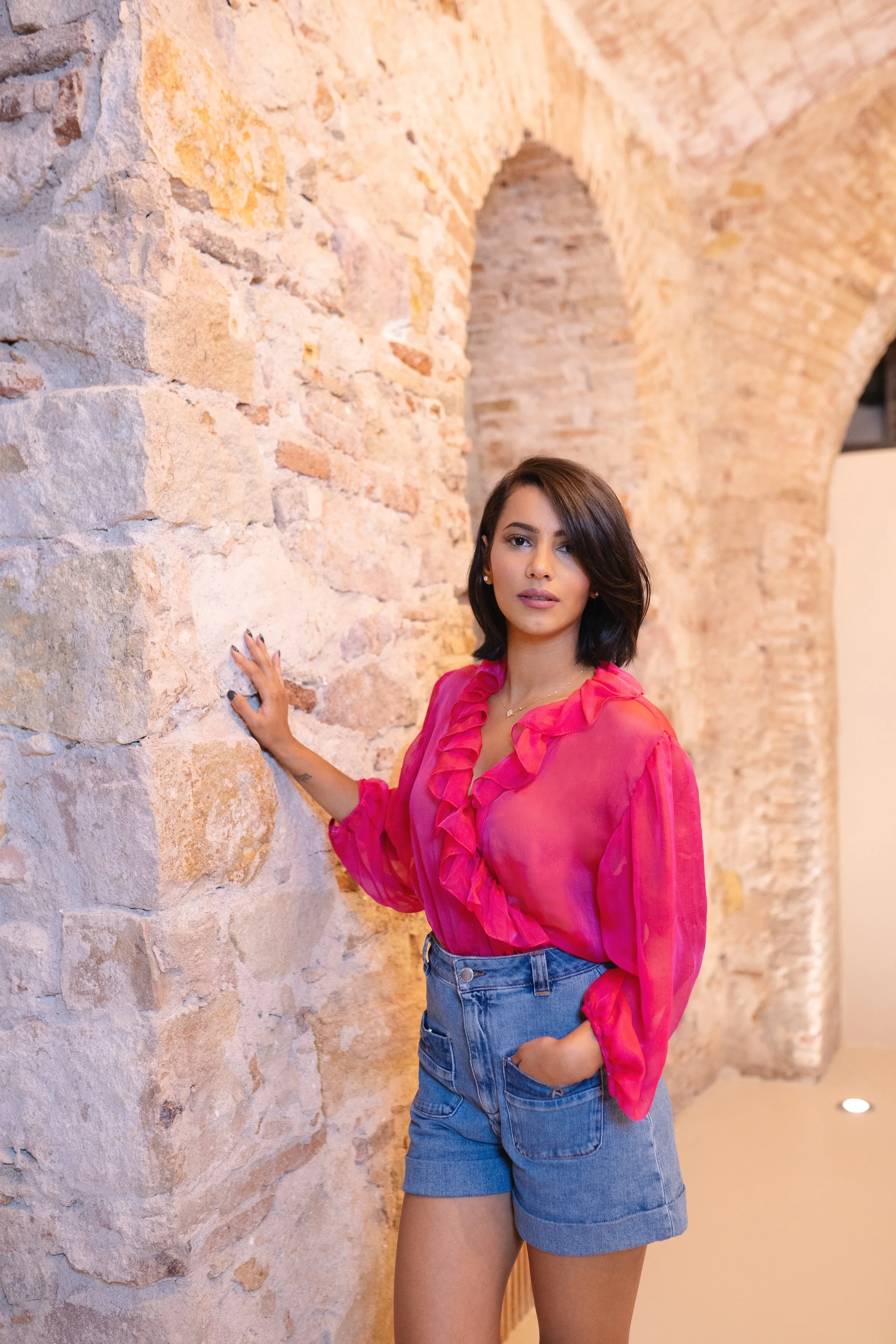 A young woman with dark hair and a pink blouse with ruffle details stands against a stone wall with an arched opening in the background, looking at the camera.