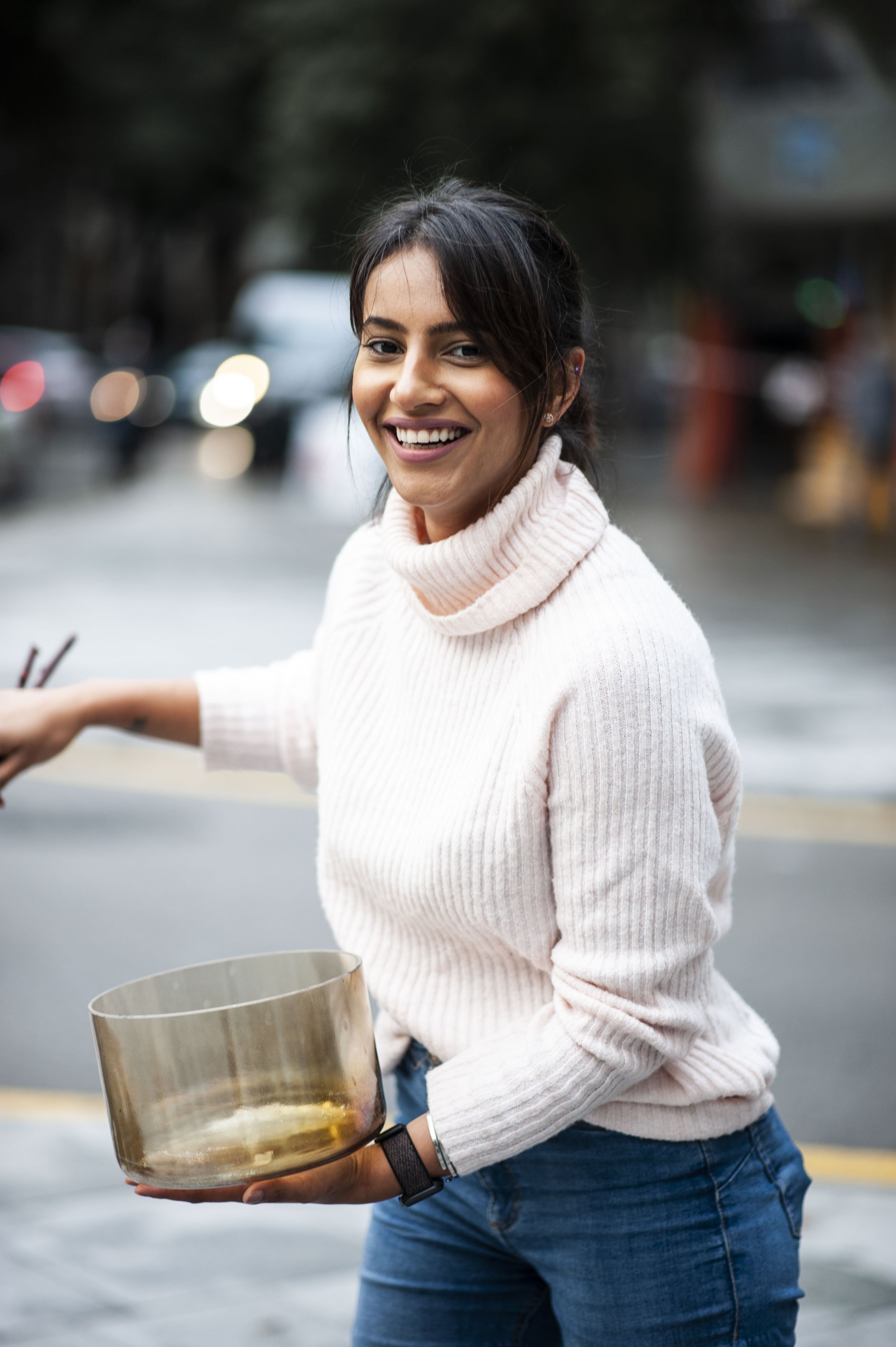 A woman smiling and holding a glass container outdoors on a city street, wearing a white turtleneck sweater and jeans.