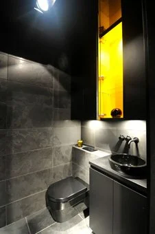 A modern bathroom with dark stone tiled walls, a wall-mounted toilet, and a black vanity with a vessel sink. There is a blackcabinet with y illuminated ellow interior and a mirror.