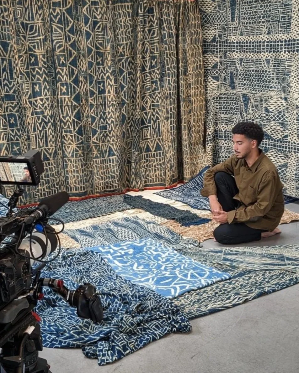 Un jeune homme assis à genoux entouré de textiles indigo aux motifs géométriques bleus et blancs, une caméra professionnelle à gauche filme la scène.