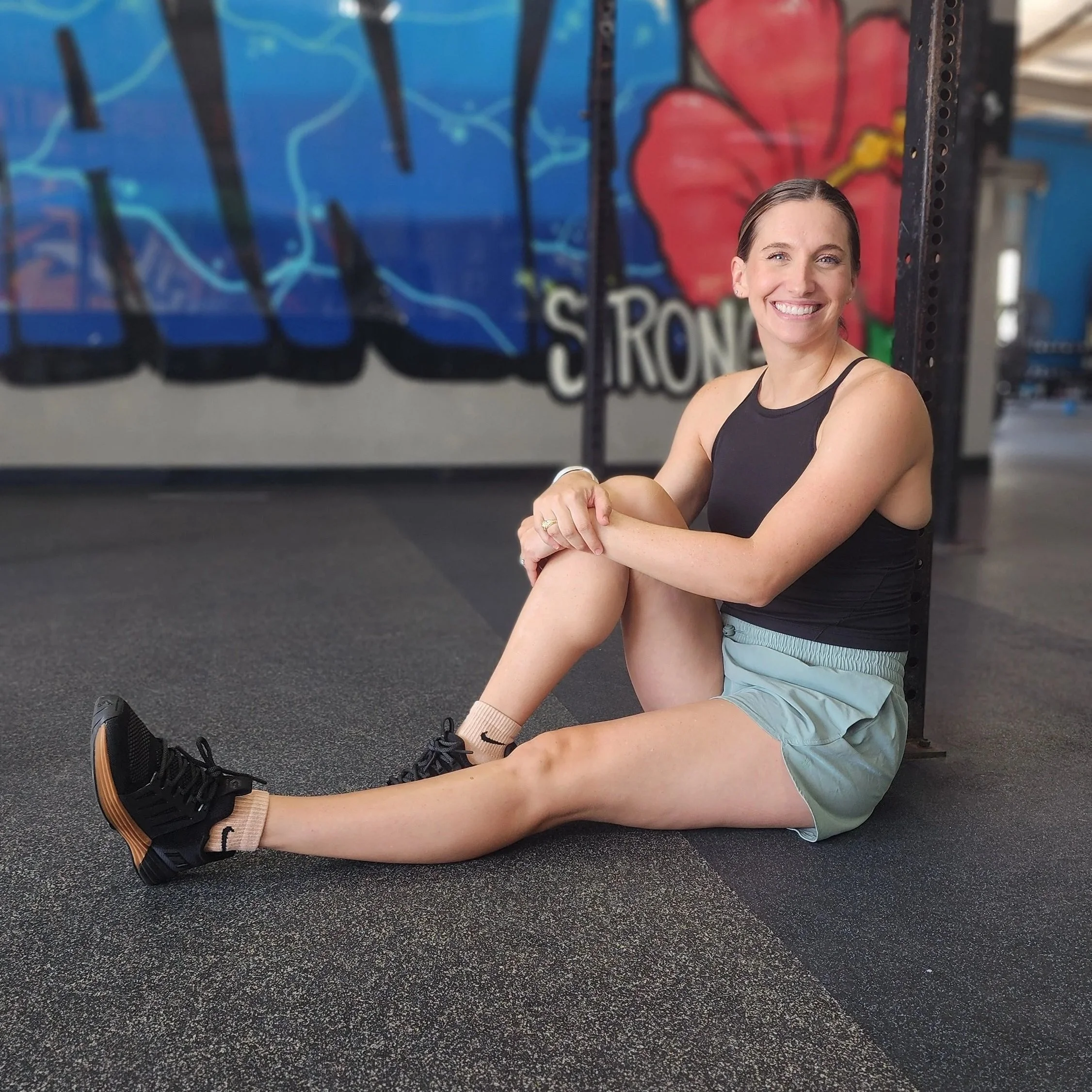 A woman smiling, sitting on the gym floor with her legs extended, in front of a colorful gym wall. She is wearing athletic clothes, including a black tank top, light-colored shorts, and black sneakers.