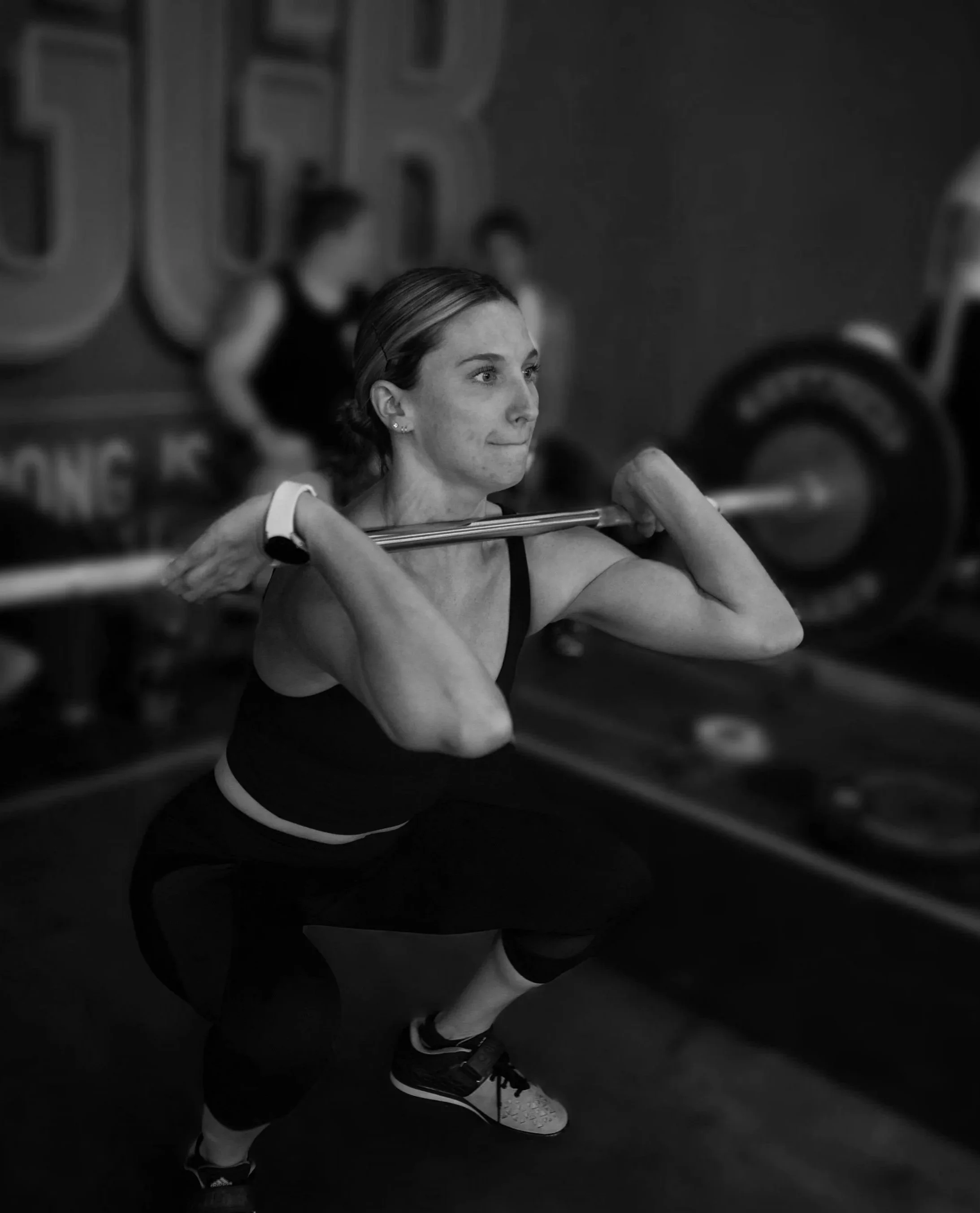 Black and white photo of a woman lifting a barbell in a gym, with a large gym logo in the background.