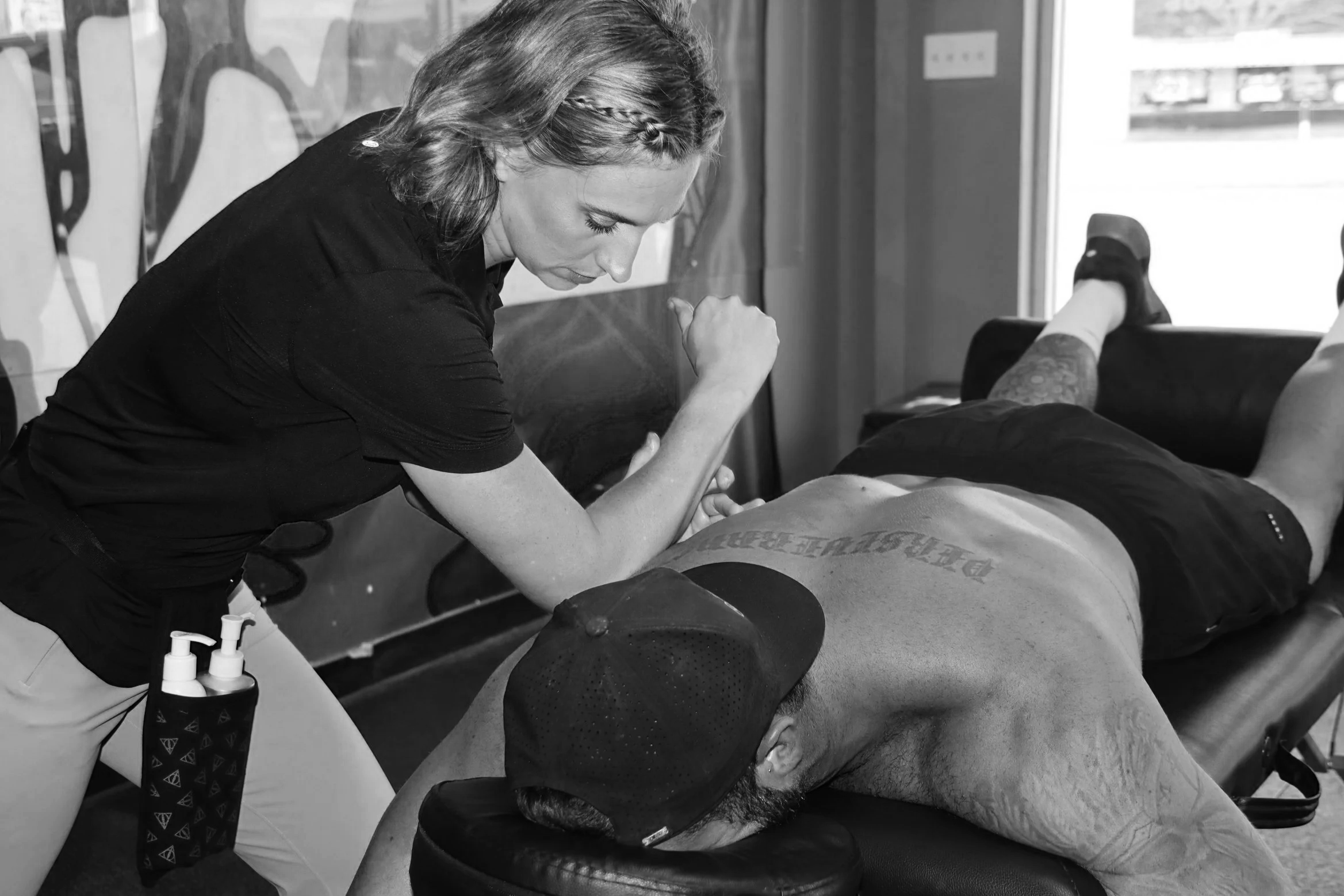 A woman in athletic clothing performs a physical therapy exercise on a man lying face down on a padded table, with her arm flexed and focused on the exercise.