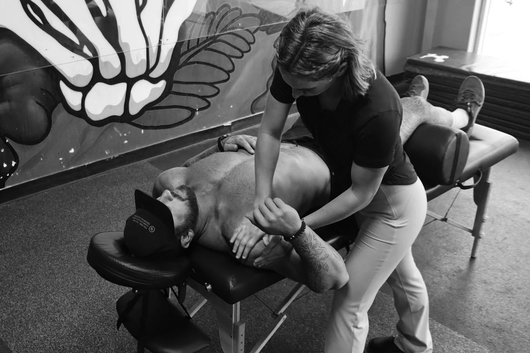 A man lying face down on a massage table getting a massage from a woman. The woman is applying pressure to his back with her hands. The setting appears to be a massage or physical therapy clinic with a decorated wall and a chair nearby.