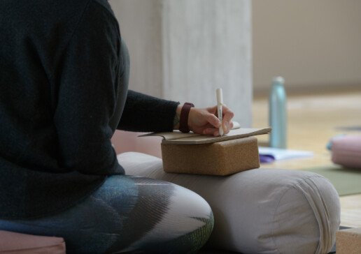 Person sitzt auf einem Meditationskissen und schreibt in ein Notizbuch in einem ruhigen Raum.