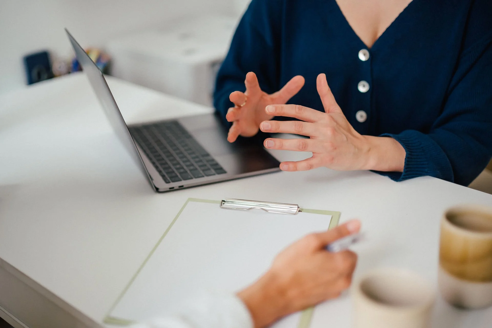 Eine Person im blauen Pullover präsentiert Gesten mit den Händen vor einem Laptop auf einem weißen Tisch, im Hintergrund sind Bürogegenstände sichtbar.