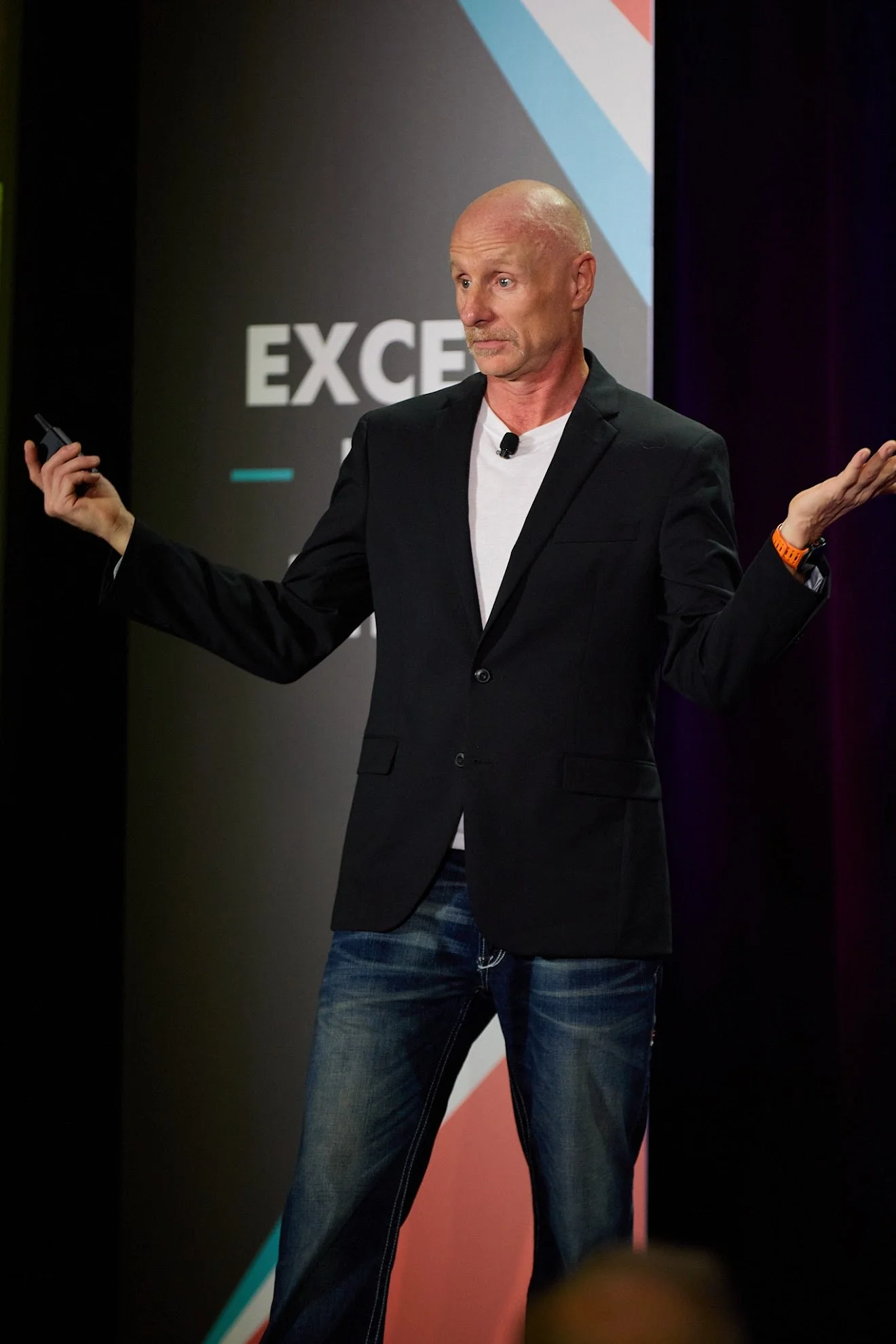 A man in a black blazer and jeans giving a presentation on stage with a background that reads "EXCE" and has a colorful geometric design.