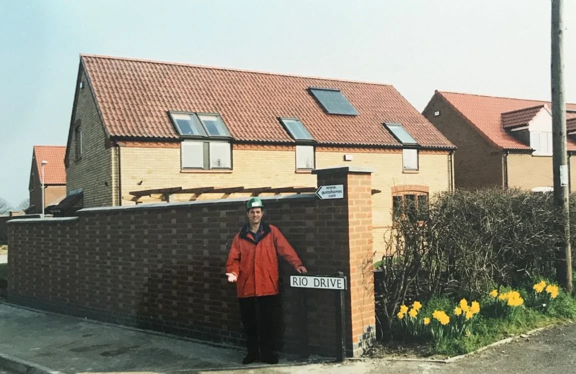 A vintage photo of Steff Wright in a red jacket and green hard hat standing next to a "Rio Drive" street sign in front of brick houses.