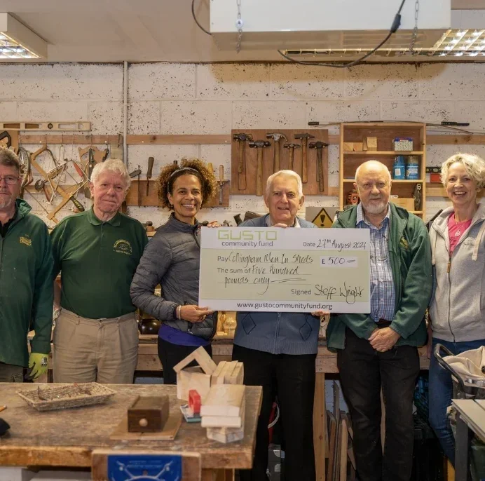 Group of six people inside a woodworking workshop, holding a large donation check for 500 GBP, with tools on the wall behind them. This is The Shed at Collingham with some members of the Gusto Community Fund. 
