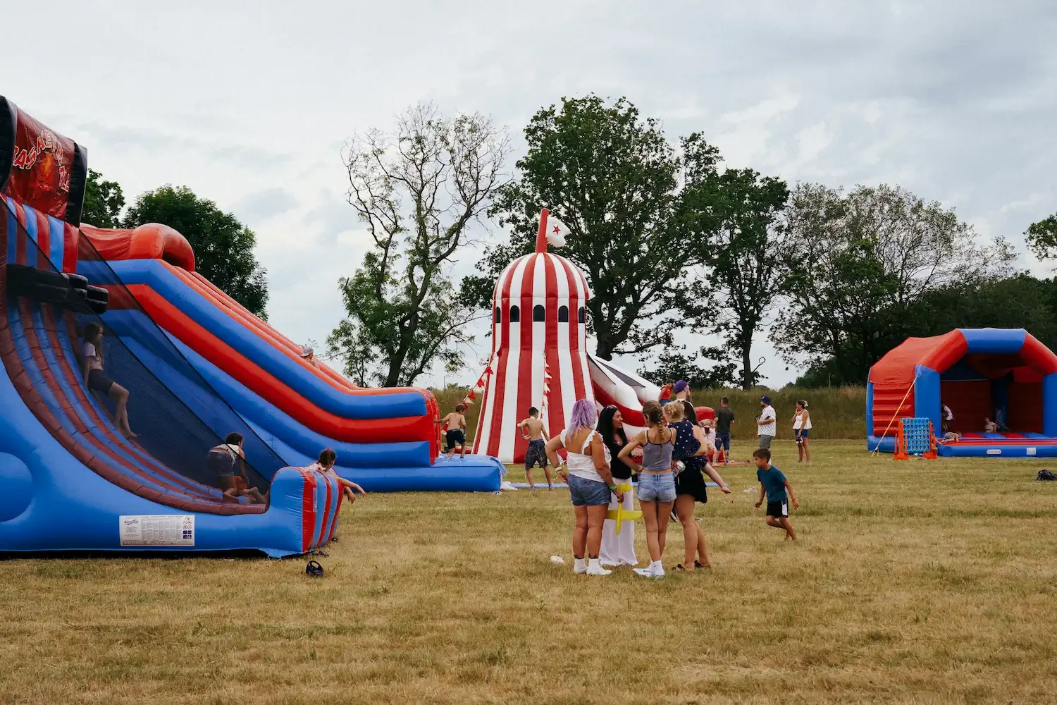 A big open green field filled with people and red, white and blue inflatables and trees in the background