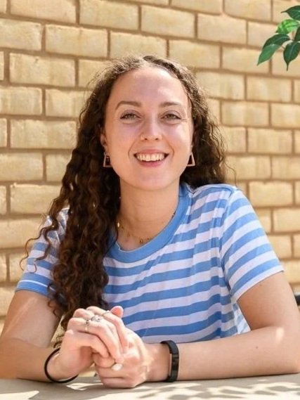 A woman with curly hair sitting at a table in front of a brick wall, smiling at the camera, wearing a blue and white striped shirt, with greenery in the background.