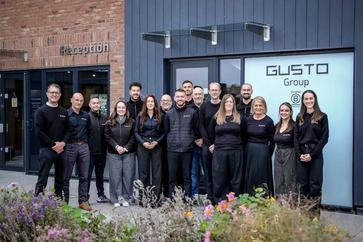 A group of approx 15 people in black gusto uniform, smiling at the camera and stood in front of a modern office building called Gusto House