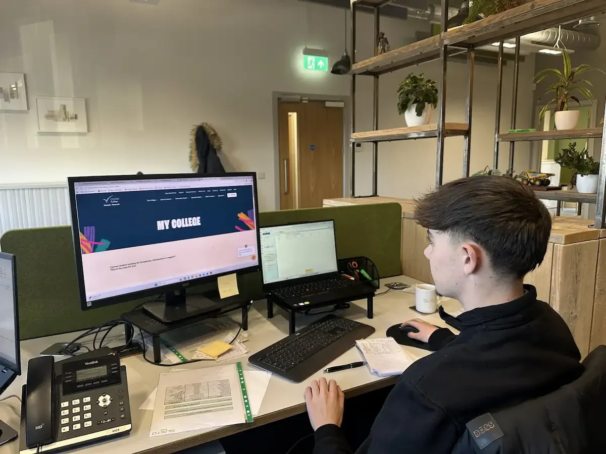 A young male sat at a desk with papers scattered everywhere, his laptop has the Lincoln College website displayed and the words 'My College'