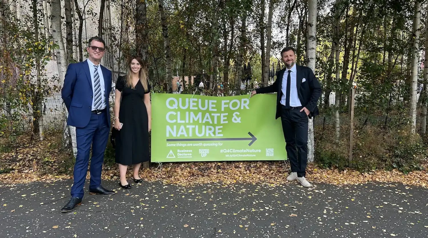 A women and two men stood infront of some trees with a green banner that reads 'Queue for Climate & Nature'