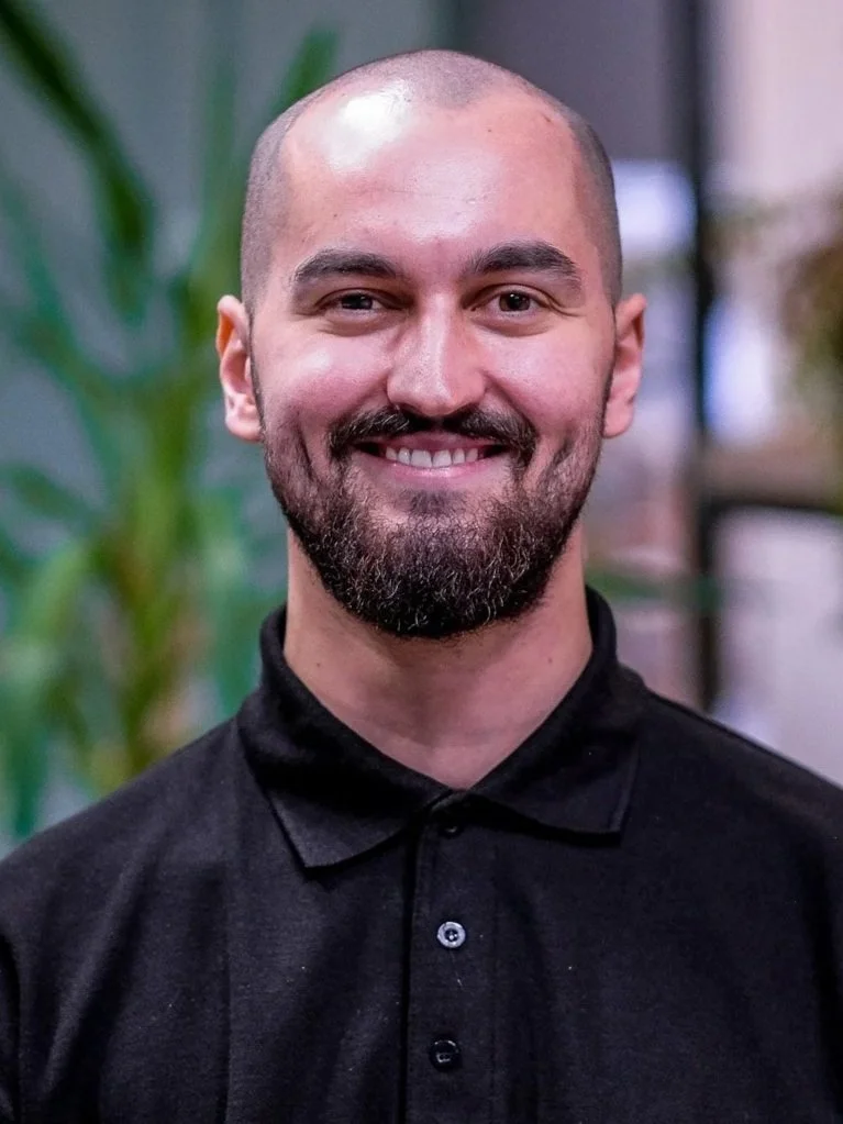 Close-up of a young man with a beard and bald head smiling in front of a blurred background.