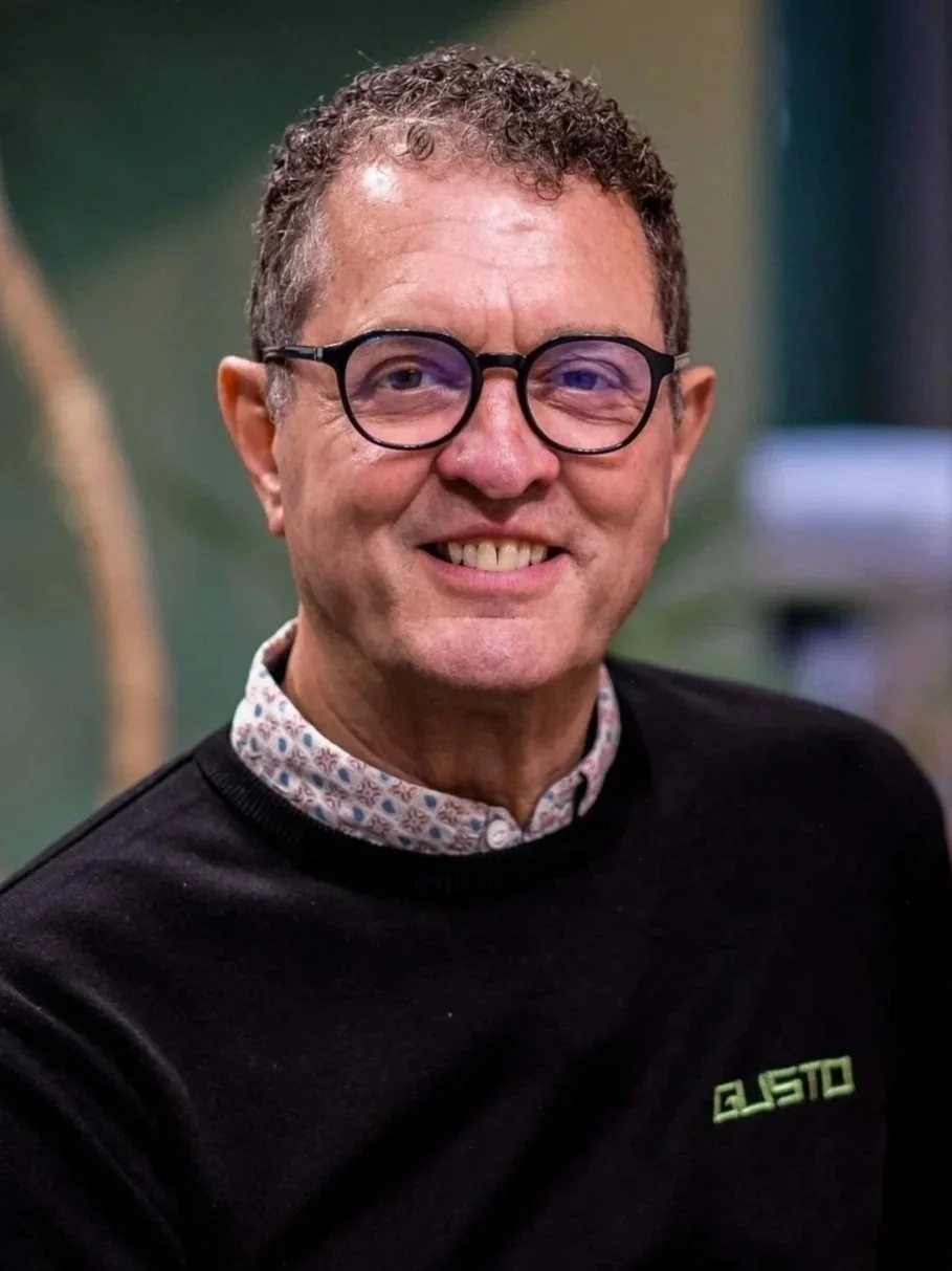 A middle-aged man with curly hair and glasses, wearing a checked shirt, standing indoors against a green and office background. Steff Wright